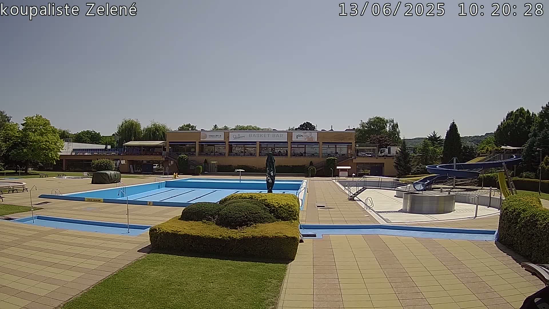 A swimming pool complex with a large pool, smaller pools, water slides, and a building under a clear, sunny sky.
