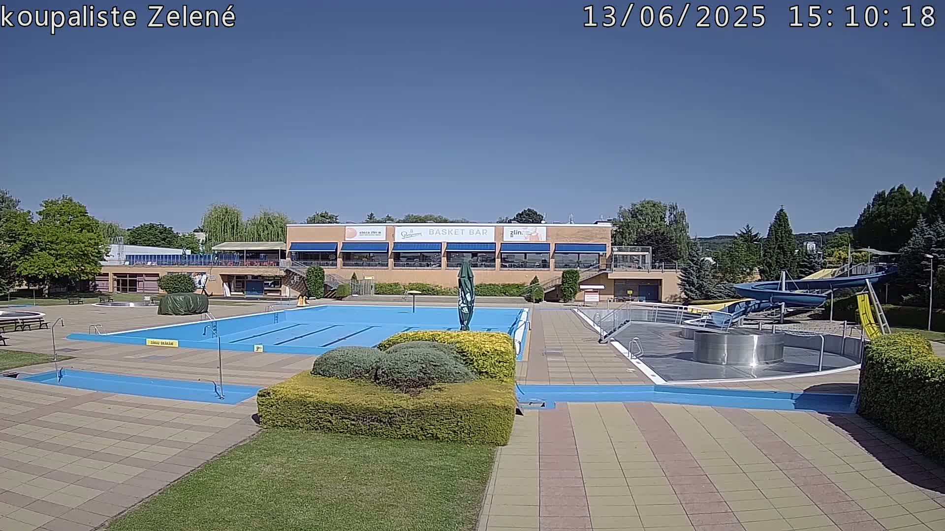 A sunny outdoor swimming pool complex with a large main pool, a smaller pool, water slides, and a building with a restaurant.