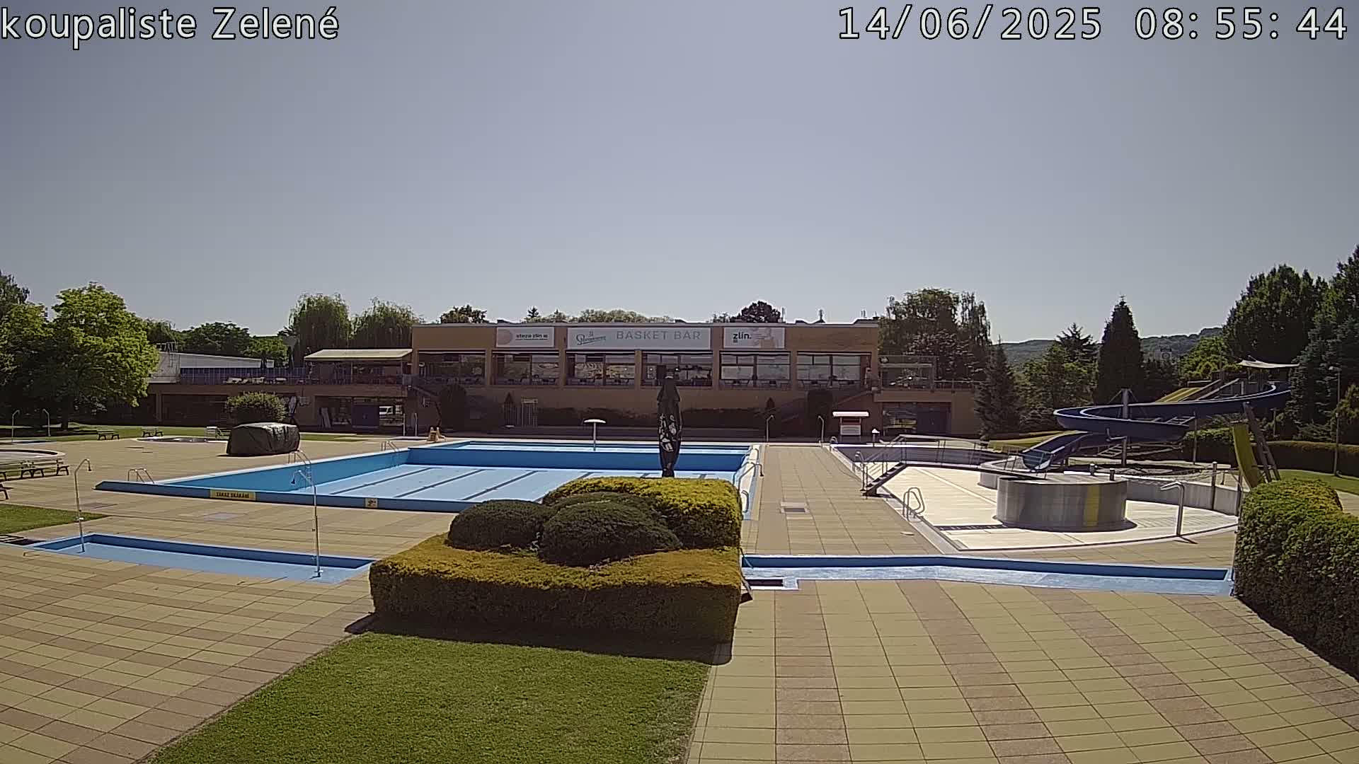 An outdoor swimming pool complex with a large main pool, smaller pools, a water slide, and a building in the background on a sunny day.