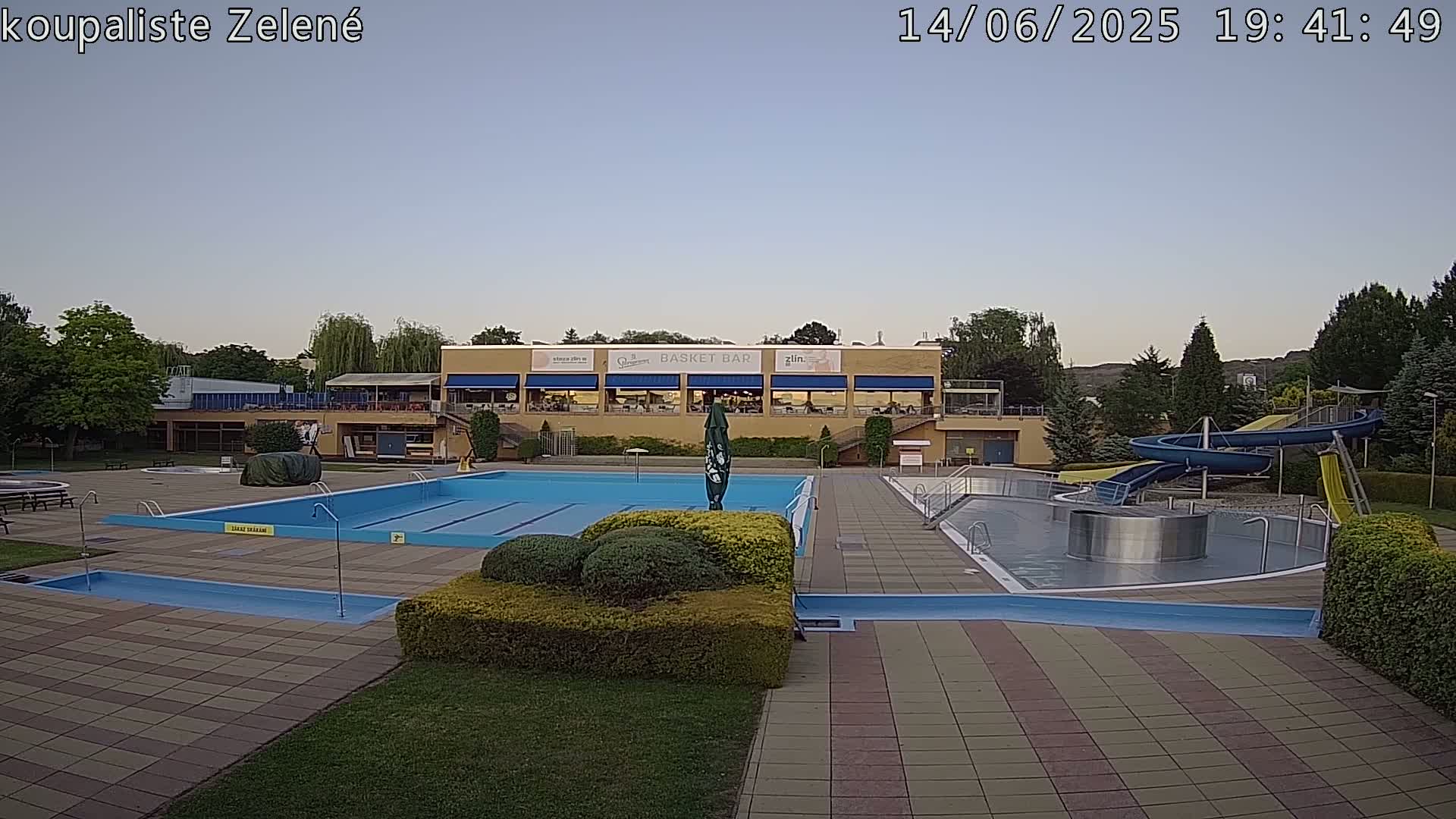 An outdoor swimming pool complex with a large main pool, a smaller wading pool, water slides, and a building with a restaurant under a clear blue sky.