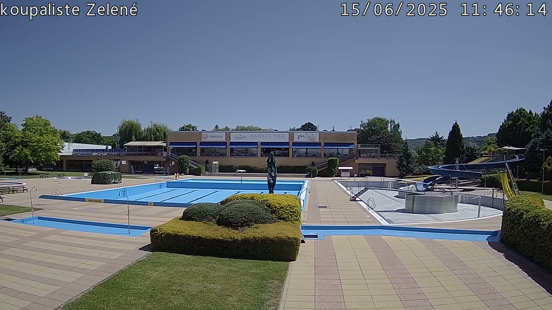 A sunny outdoor swimming pool complex with a large main pool, smaller wading pool, water slides, and a building with a restaurant.