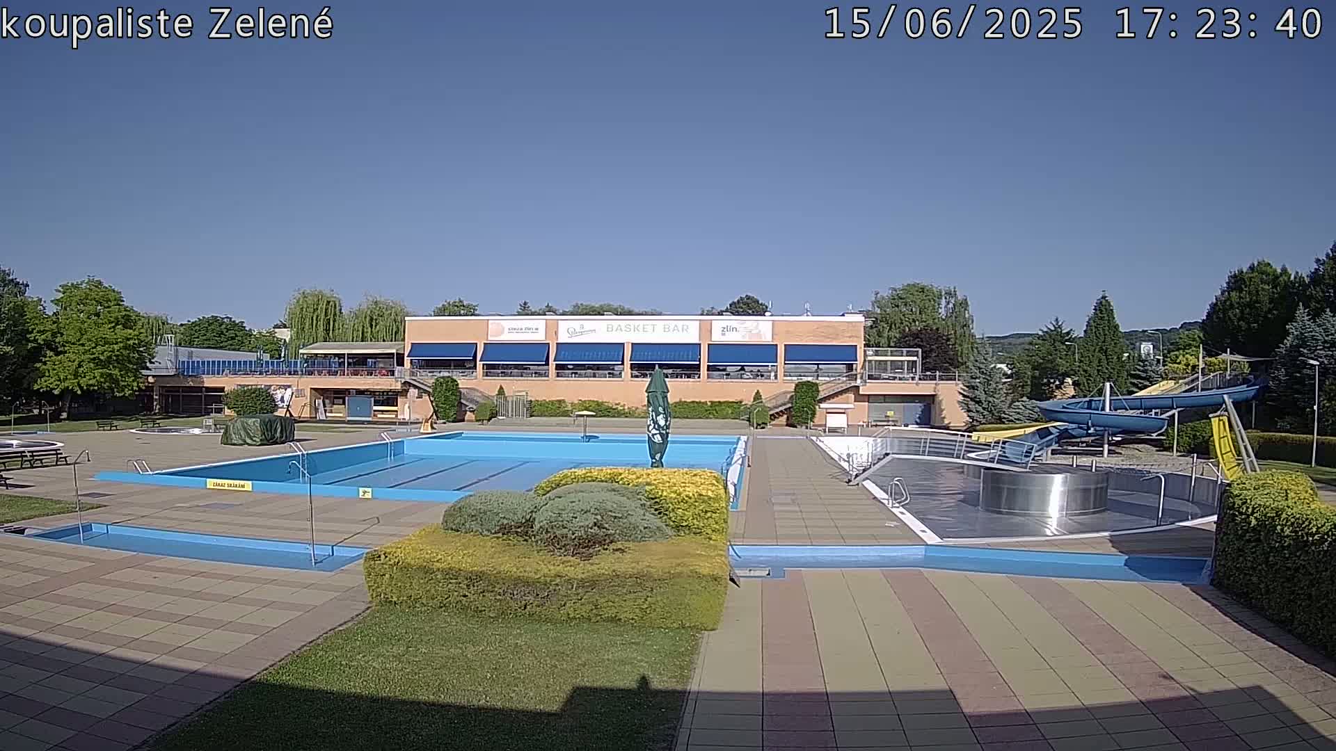 An outdoor pool complex on a sunny day features a large rectangular pool, a smaller pool, water slides, and a building with a snack bar.