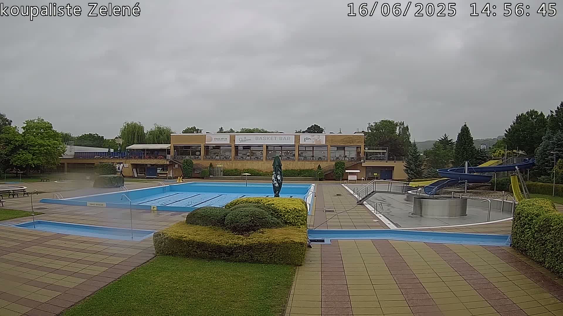 An outdoor swimming pool complex with a large rectangular pool, smaller wading pool, water slides, and a building with a restaurant under an overcast sky.