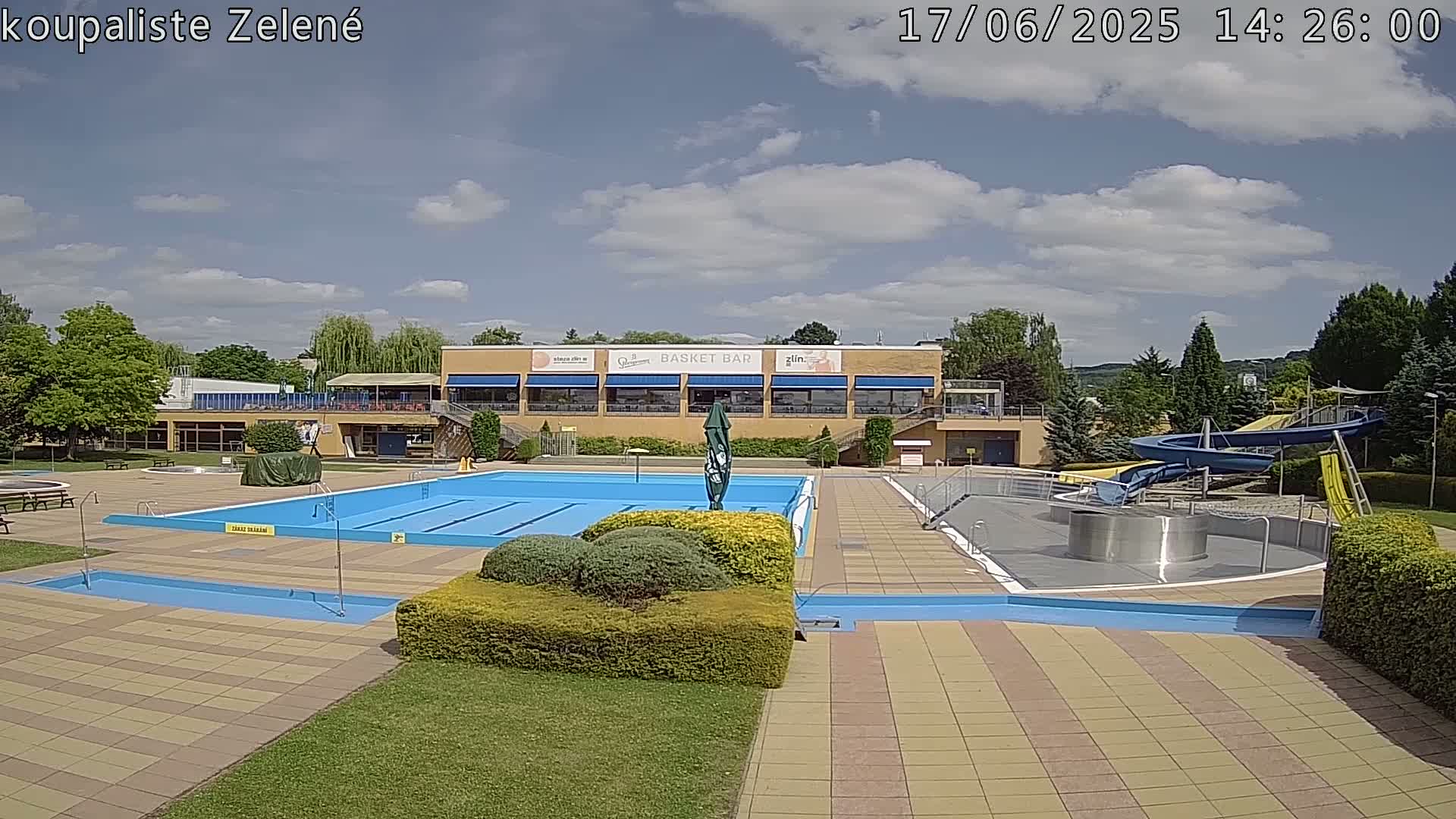 An outdoor pool complex with a large swimming pool, smaller pools, water slides, and a building with a restaurant under partly cloudy skies.