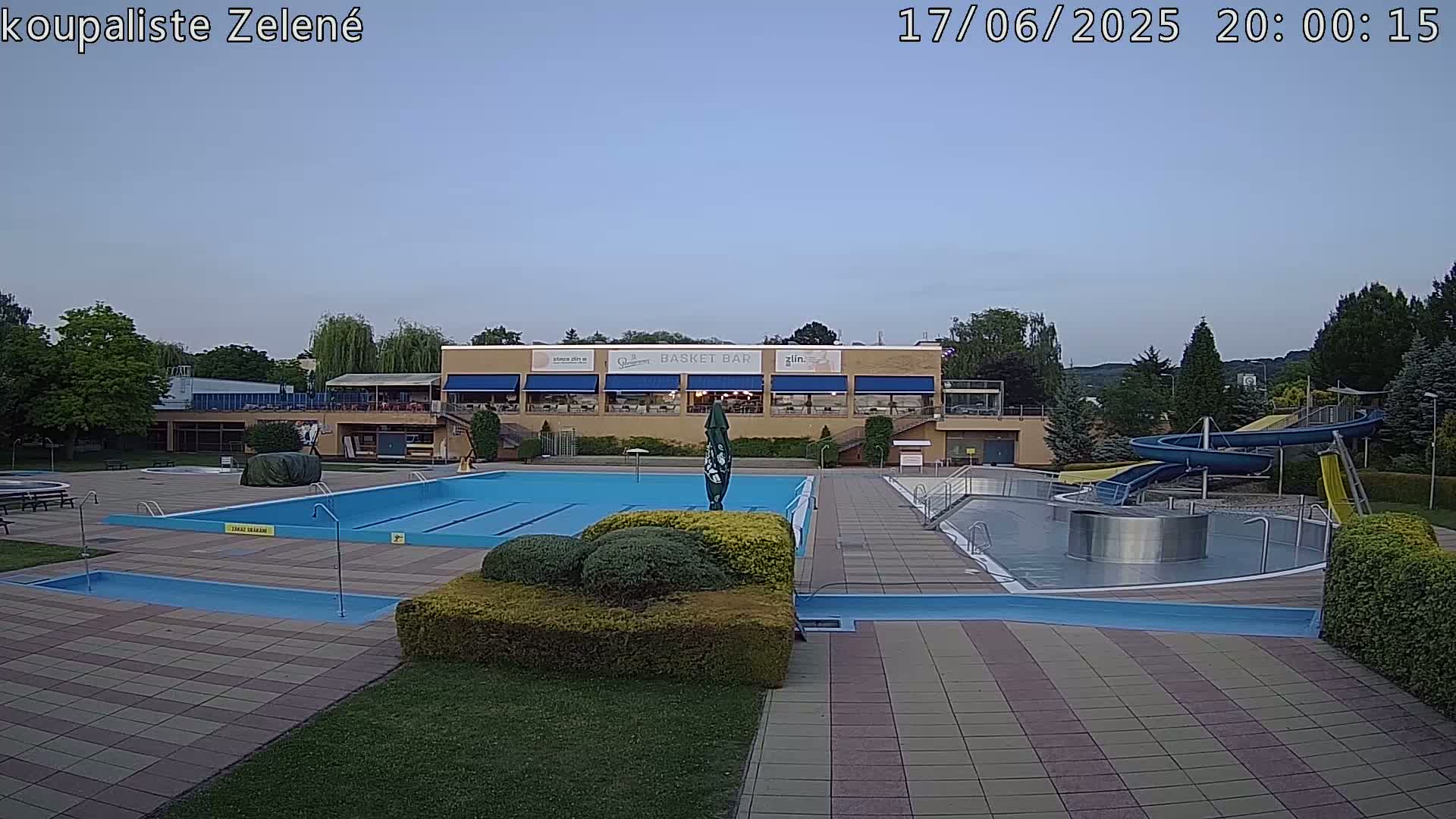 An outdoor pool complex with a large swimming pool, a smaller wading pool, water slides, and a building with a restaurant.  The sky is clear and light blue.