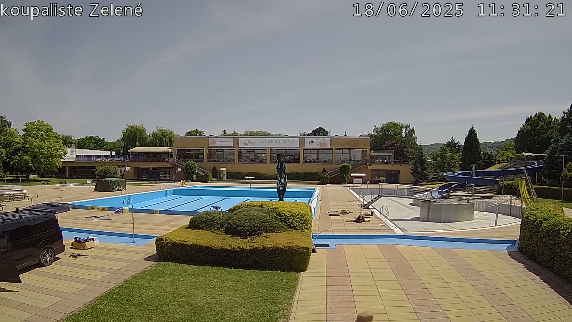An outdoor swimming pool complex with a large swimming pool, water slides, and a building in the background under a mostly sunny sky.