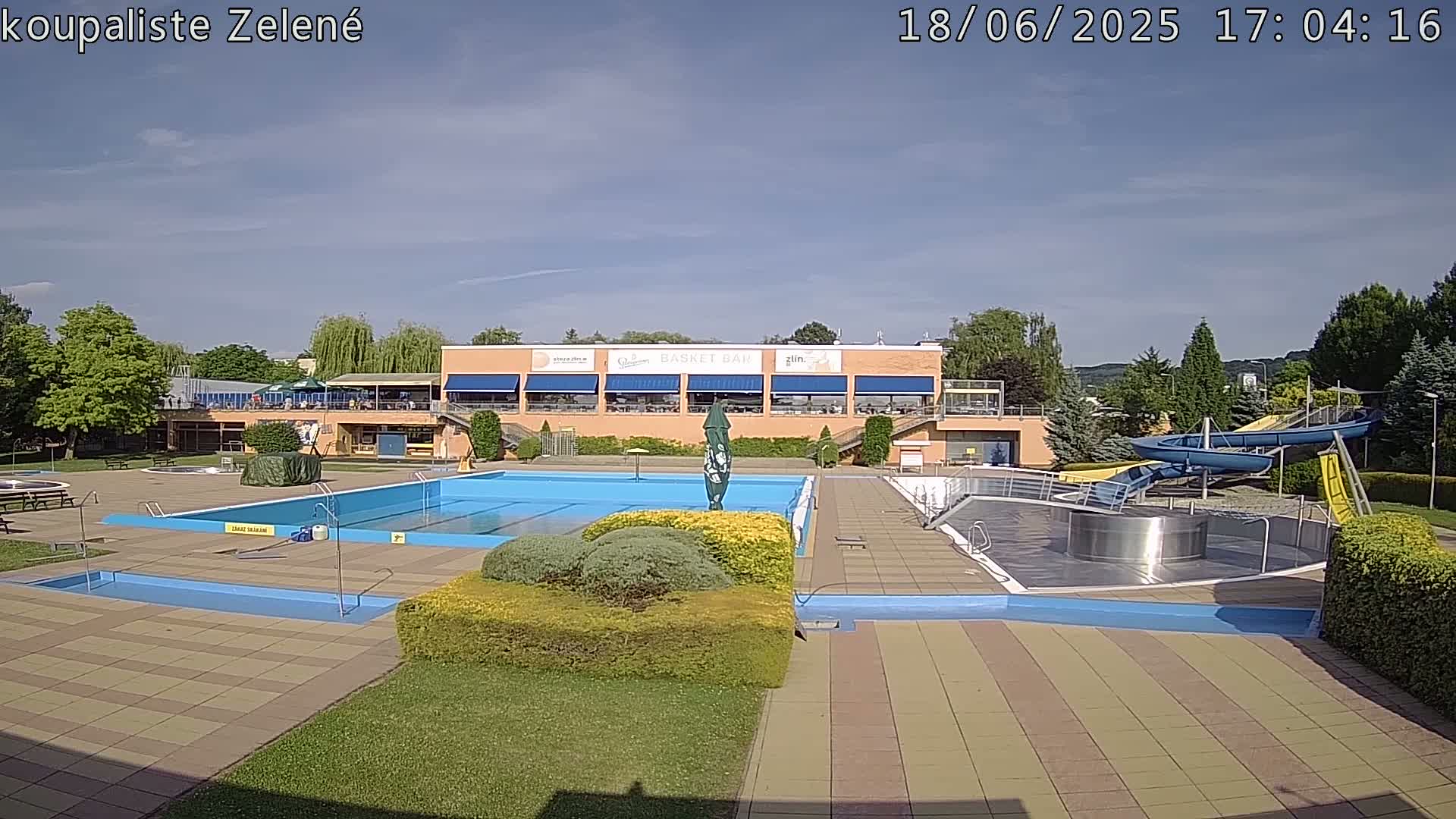 An outdoor swimming pool complex with a large main pool, smaller pools, water slides, and a building with a restaurant area under mostly sunny skies.