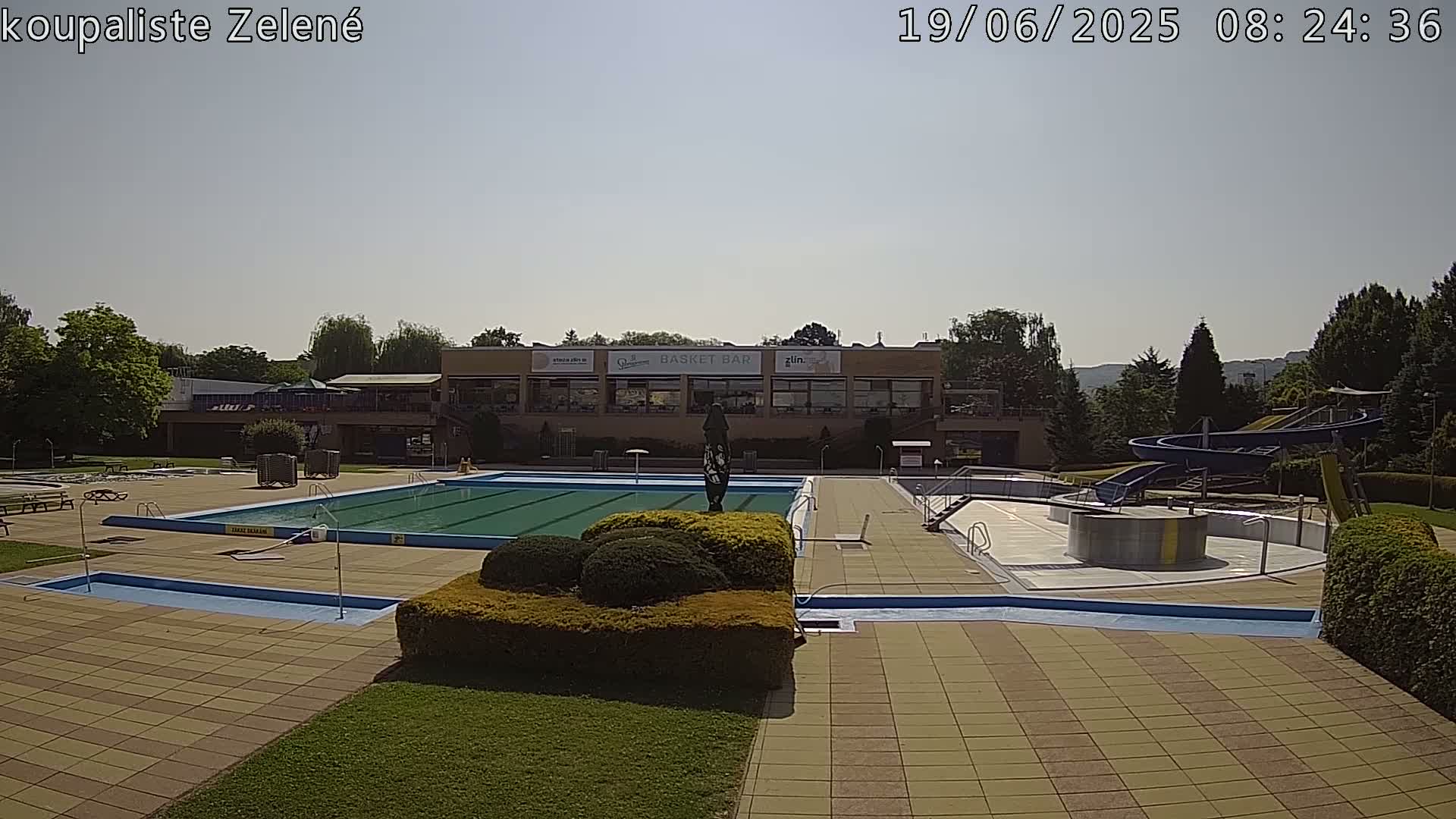An outdoor pool area with a large swimming pool, water slides, and a building in the background on a sunny day.