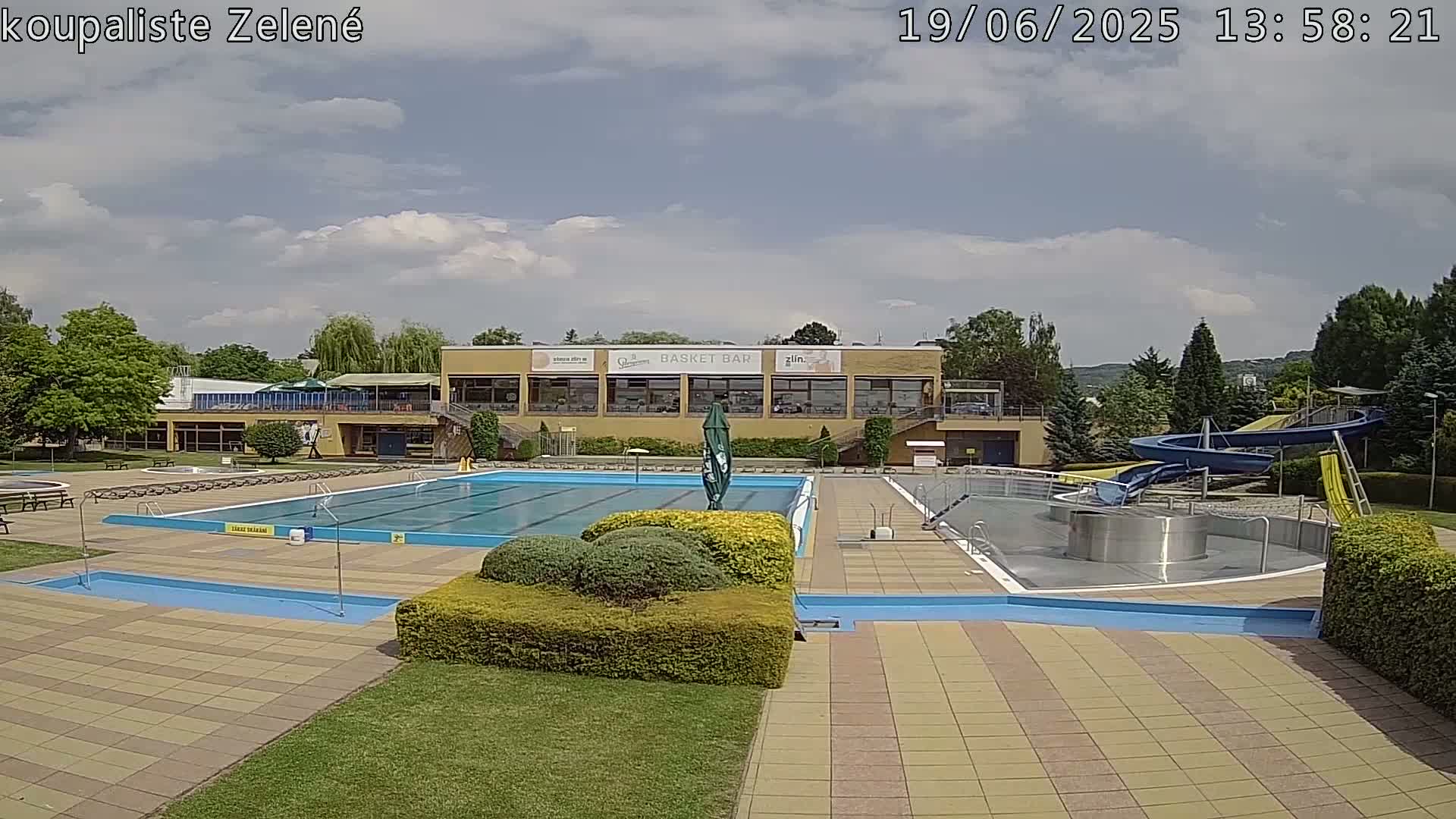 A sunny outdoor swimming complex with a large pool, a water slide, and a building with a restaurant.
