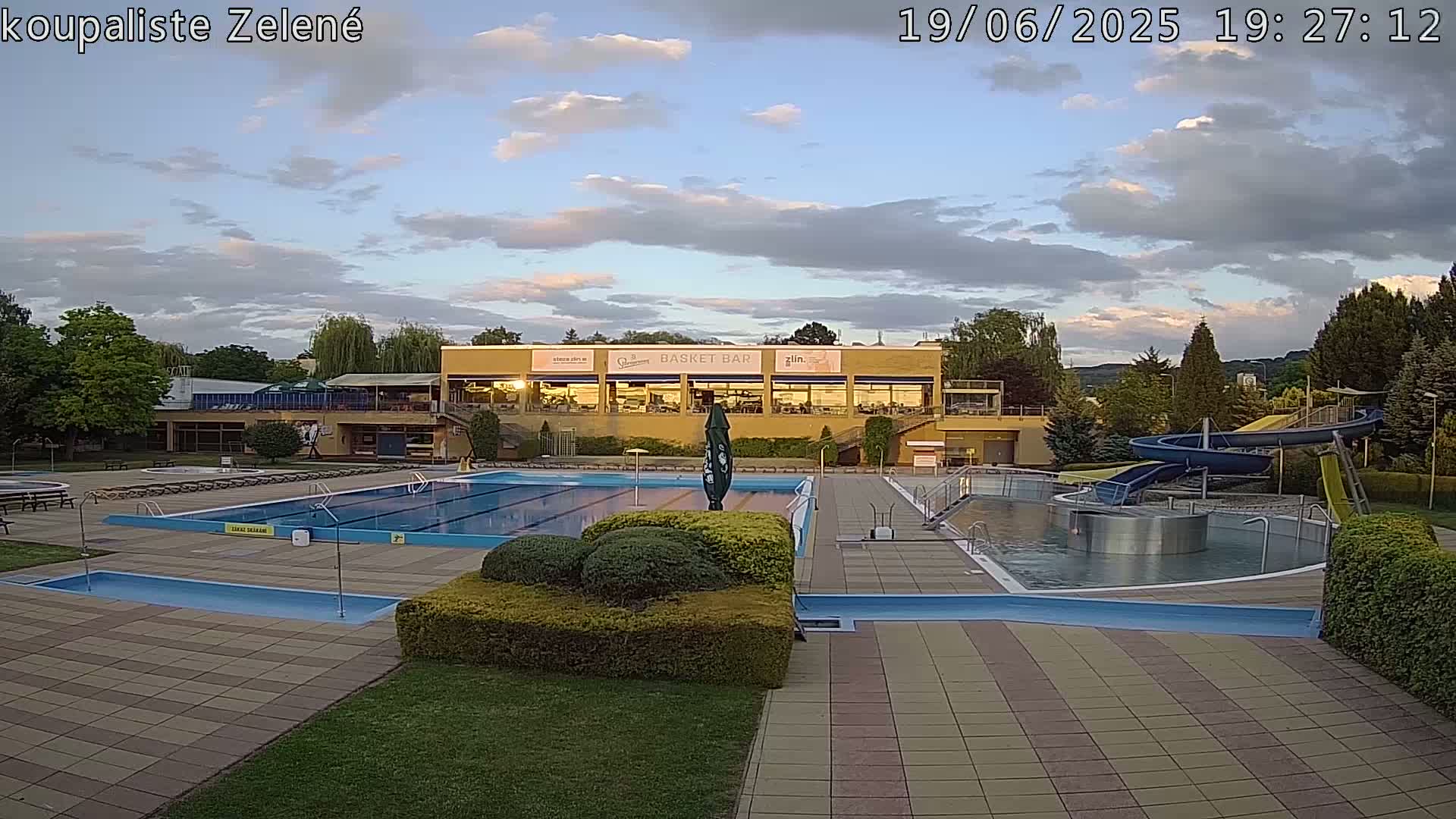 An outdoor swimming pool complex with a water slide, under a partly cloudy sky.