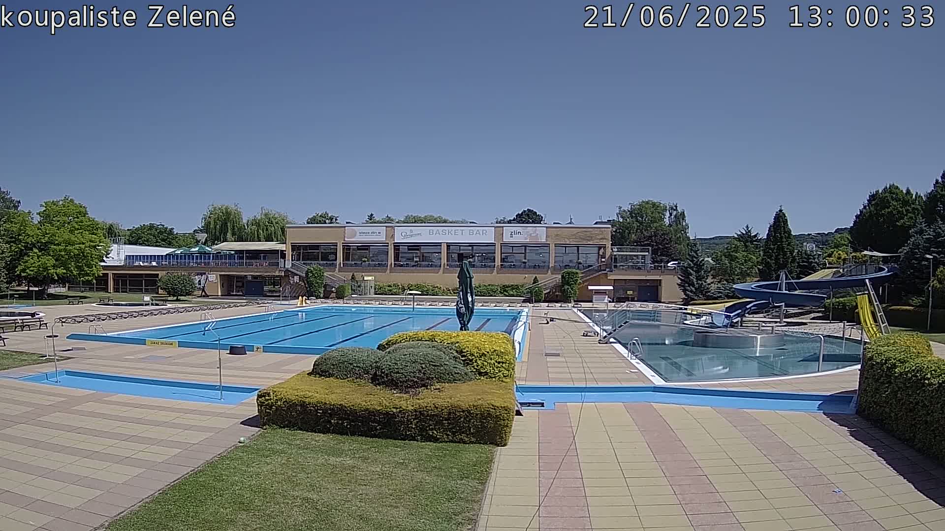 A sunny outdoor scene shows a large swimming pool complex with a water slide, surrounded by landscaping and a building with a restaurant.