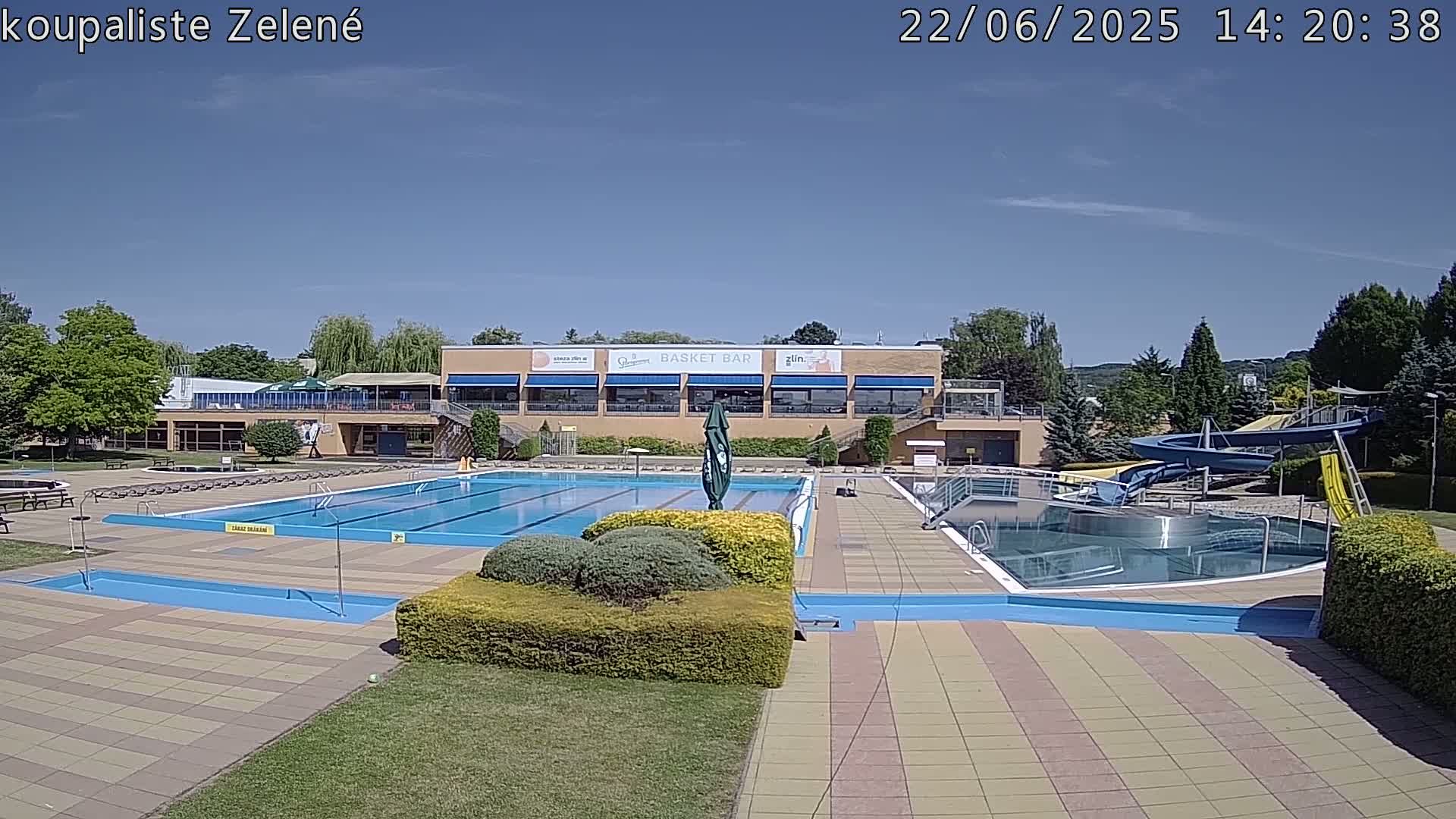 A sunny outdoor scene shows a large swimming pool complex with multiple pools, water slides, and a building with a snack bar.