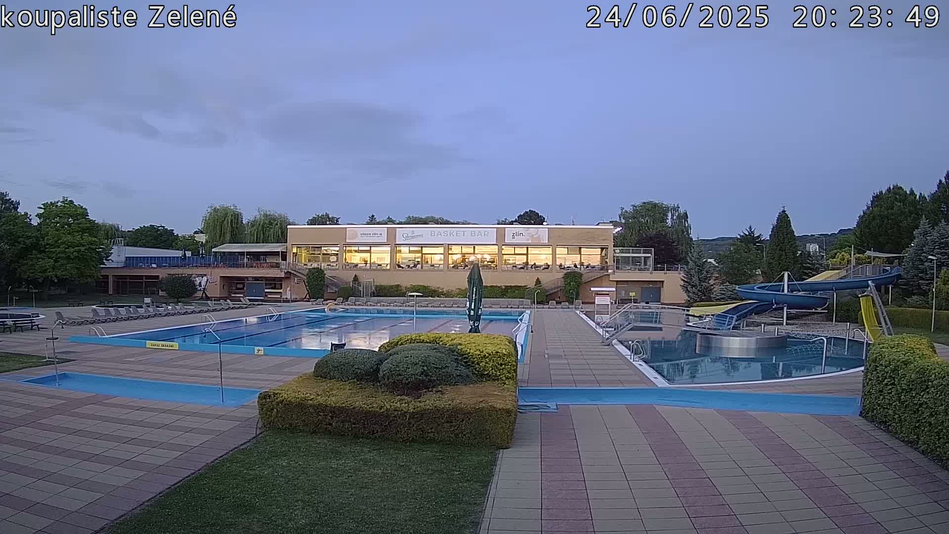 An outdoor swimming pool complex at dusk, featuring a large rectangular pool, a smaller circular pool with water slides, and a building with illuminated windows in the background.