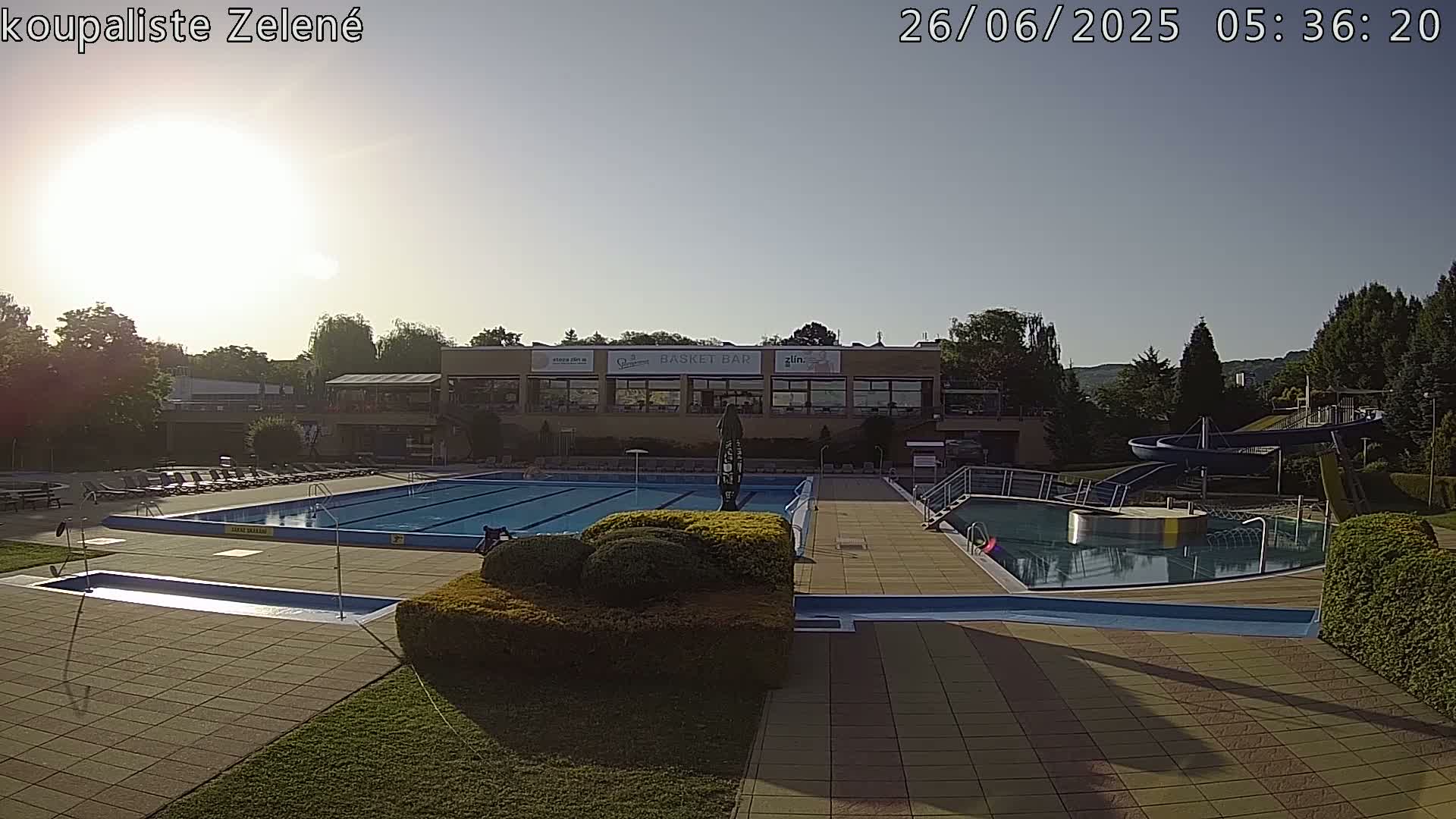 An outdoor swimming pool complex at dusk, featuring a large rectangular pool, a smaller circular pool with water slides, and a building with illuminated windows in the background.