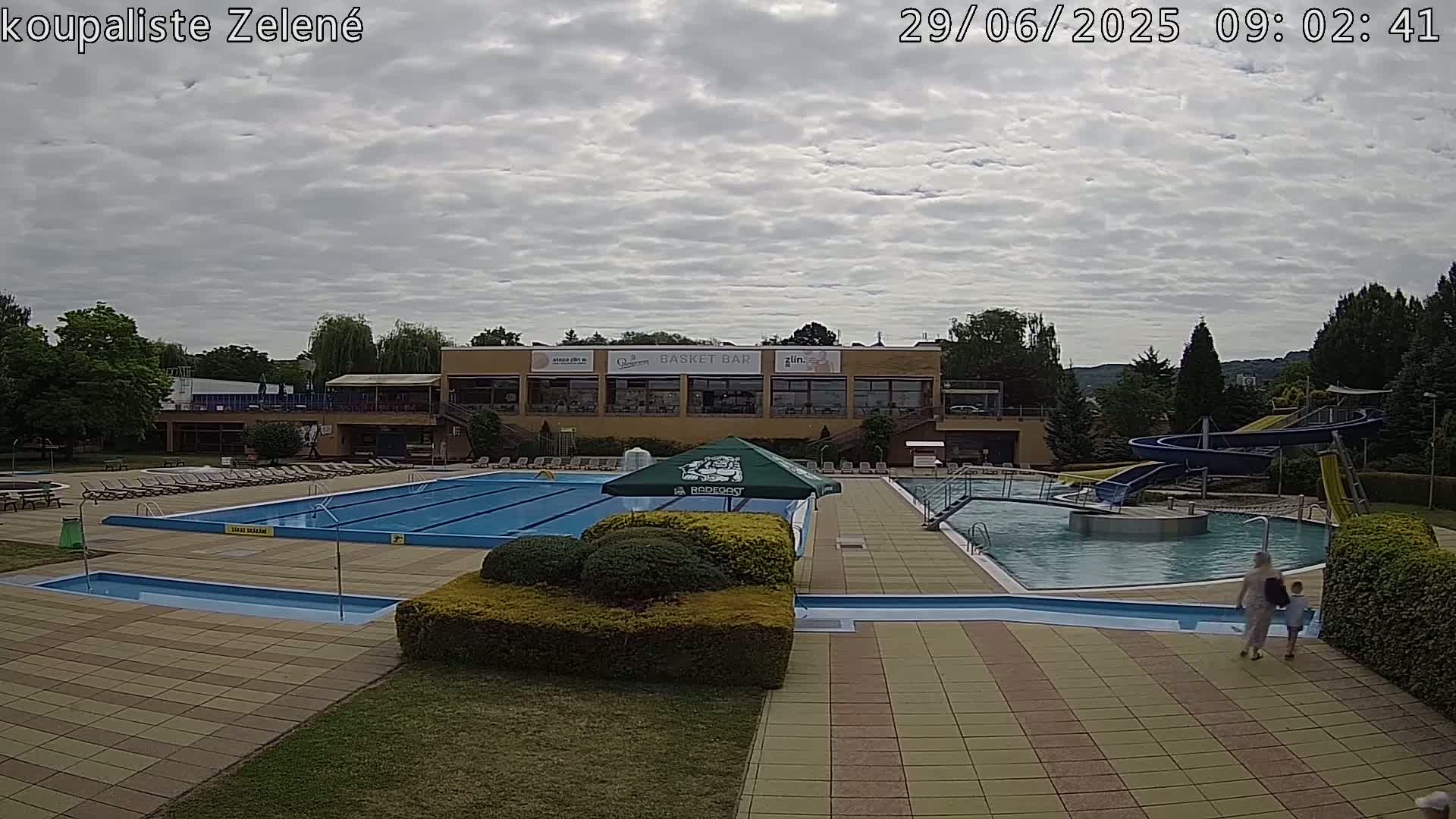 An outdoor pool complex with a large swimming pool, smaller pools, water slides, and lounge chairs under a cloudy sky.