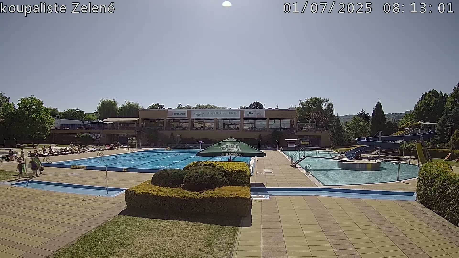 A sunny outdoor swimming pool complex with a large main pool, a smaller pool with water slides, and several people relaxing on lounge chairs.