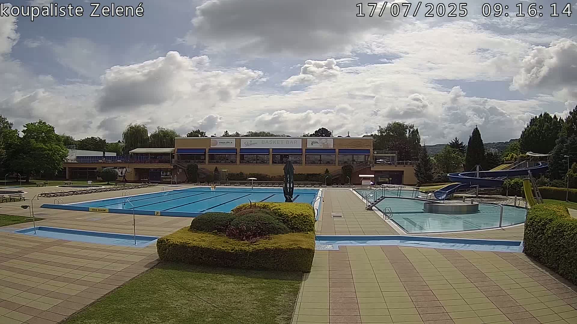 An outdoor pool complex with a large swimming pool, a smaller pool, water slides, and a building with a patio under a partly cloudy sky.