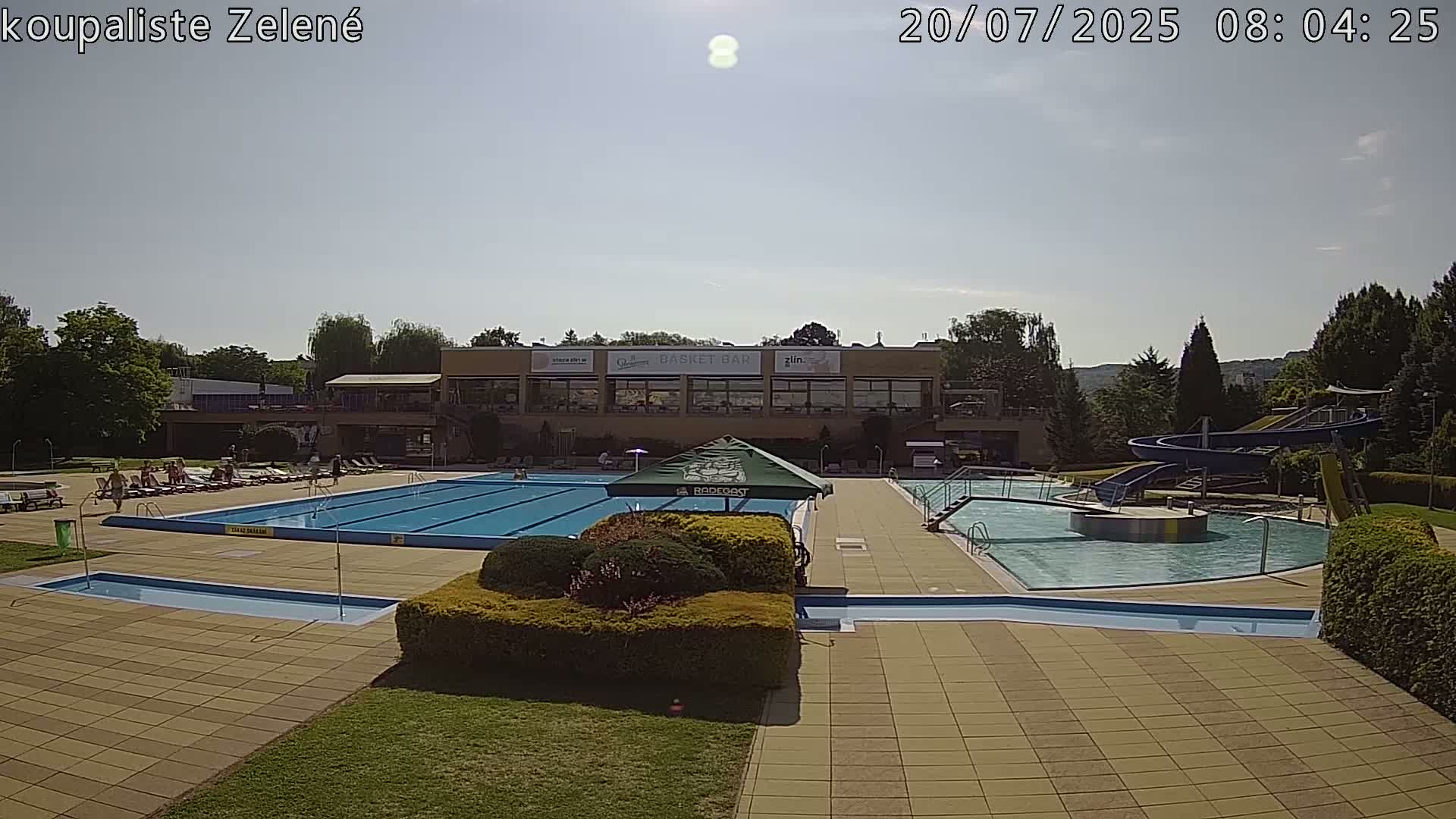 A sunny outdoor swimming complex features a large main pool, a smaller pool with a water slide, and lounge chairs surrounding the pools.