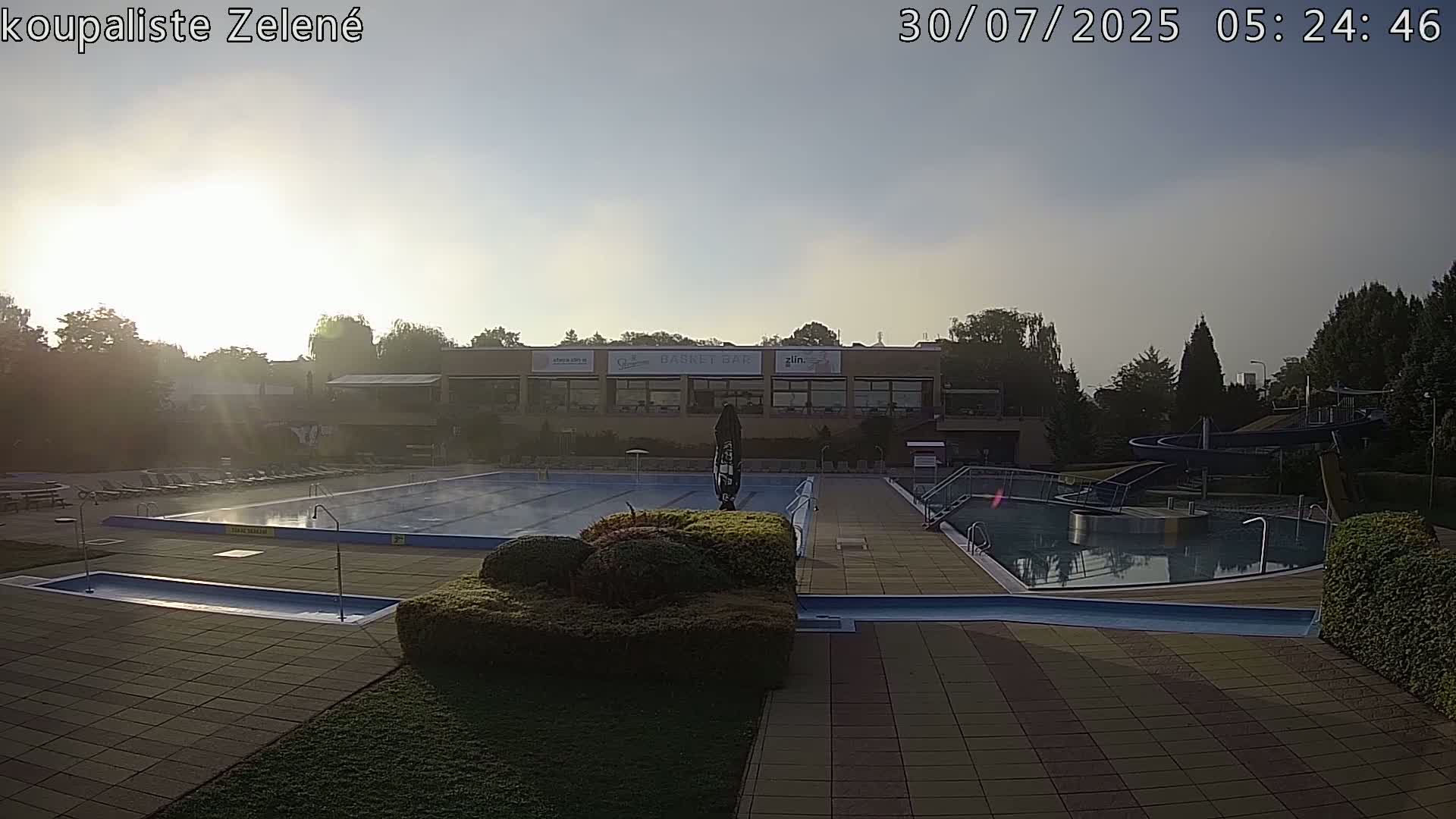 A sunny outdoor swimming complex features a large main pool, a smaller pool with a water slide, and lounge chairs surrounding the pools.
