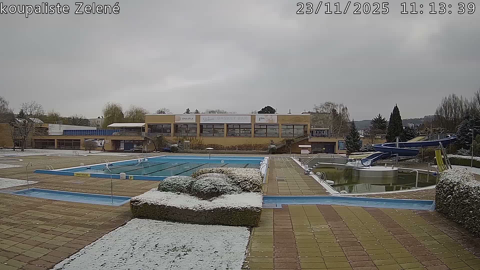 An empty outdoor swimming pool complex, lightly dusted with snow under an overcast sky, features multiple pools (some filled, one murky), a water slide, and a main building.