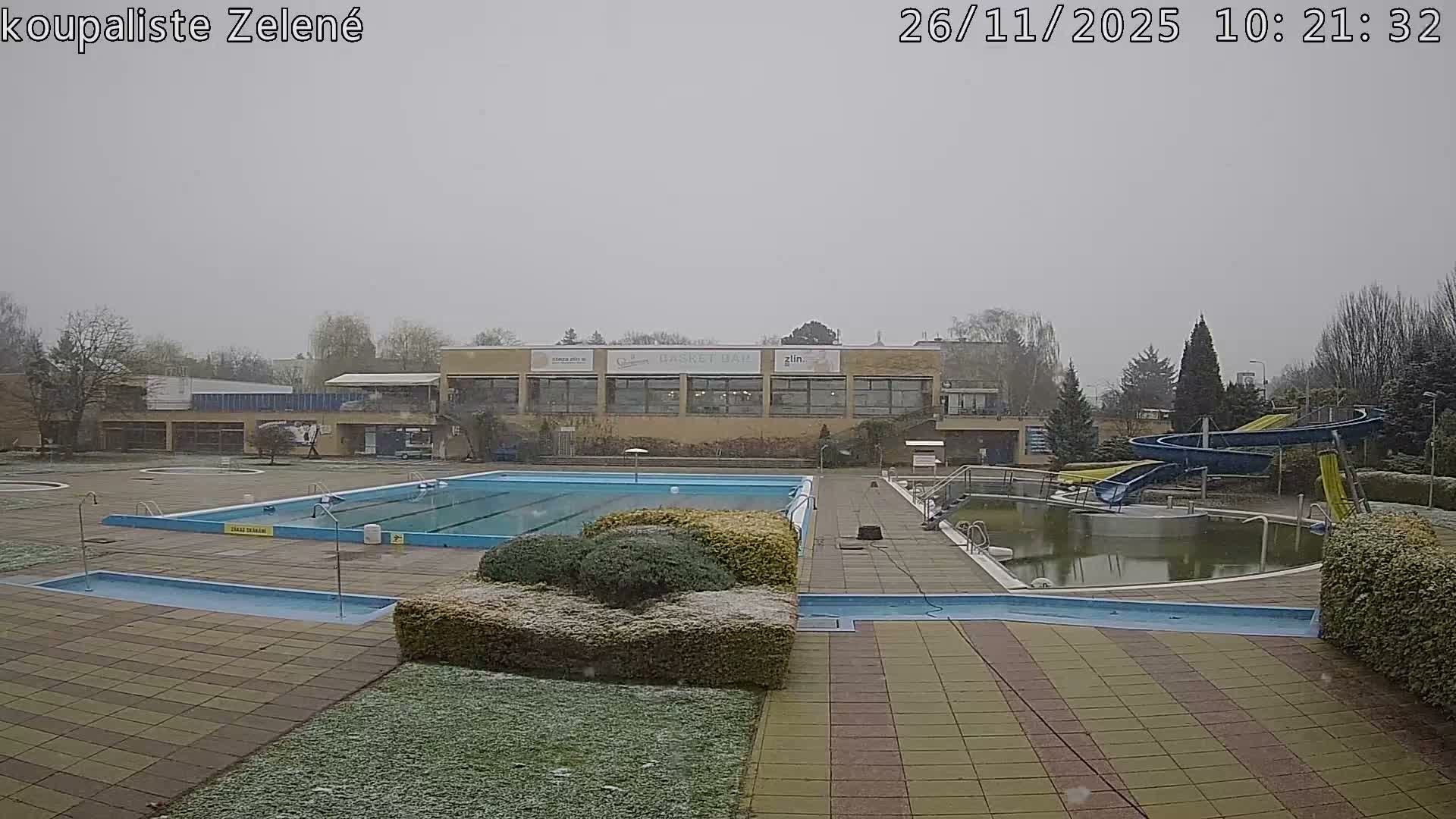 An empty outdoor swimming pool complex is depicted on an overcast day with light snow falling, showing multiple pools, some murky and some blue, surrounded by tiled pavement, snow-dusted bushes, and a building with water slides in the background.