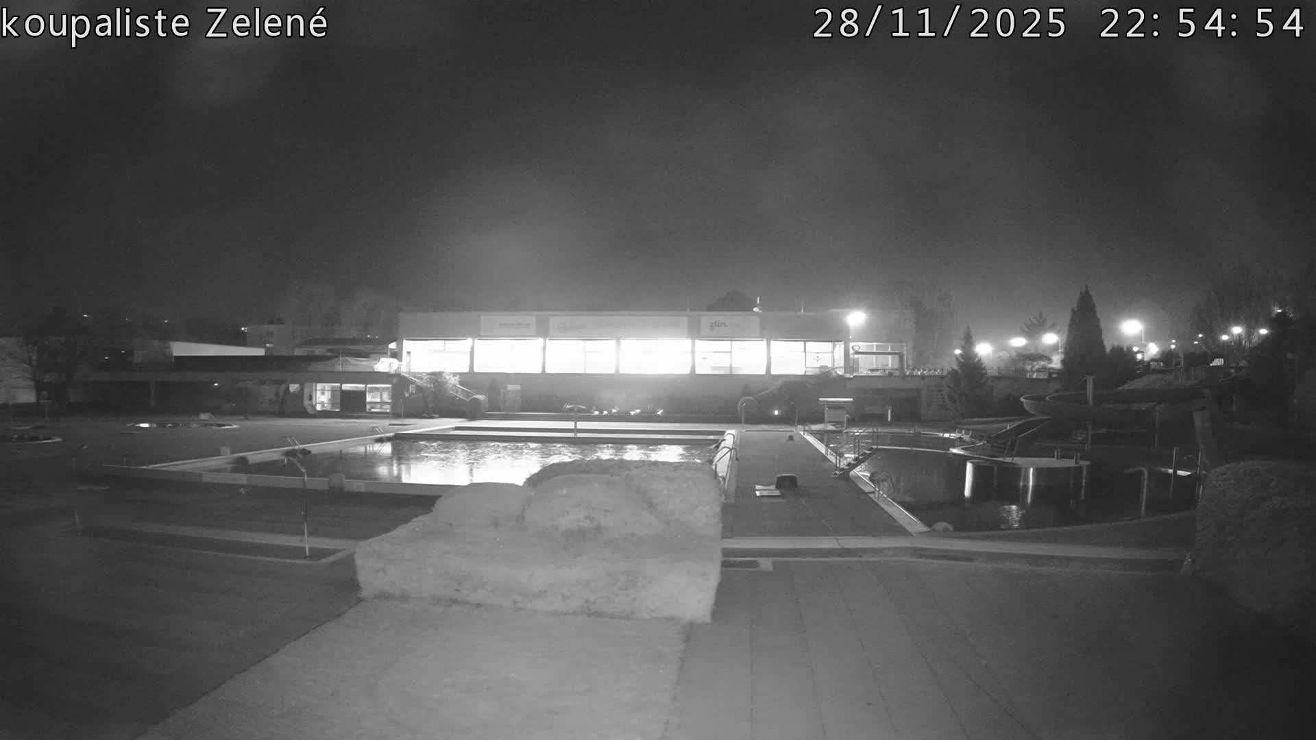 An outdoor swimming facility, featuring illuminated buildings, multiple pools, water slides, and trees, is seen at night under foggy conditions, with a significant snowdrift covering a section of the foreground.