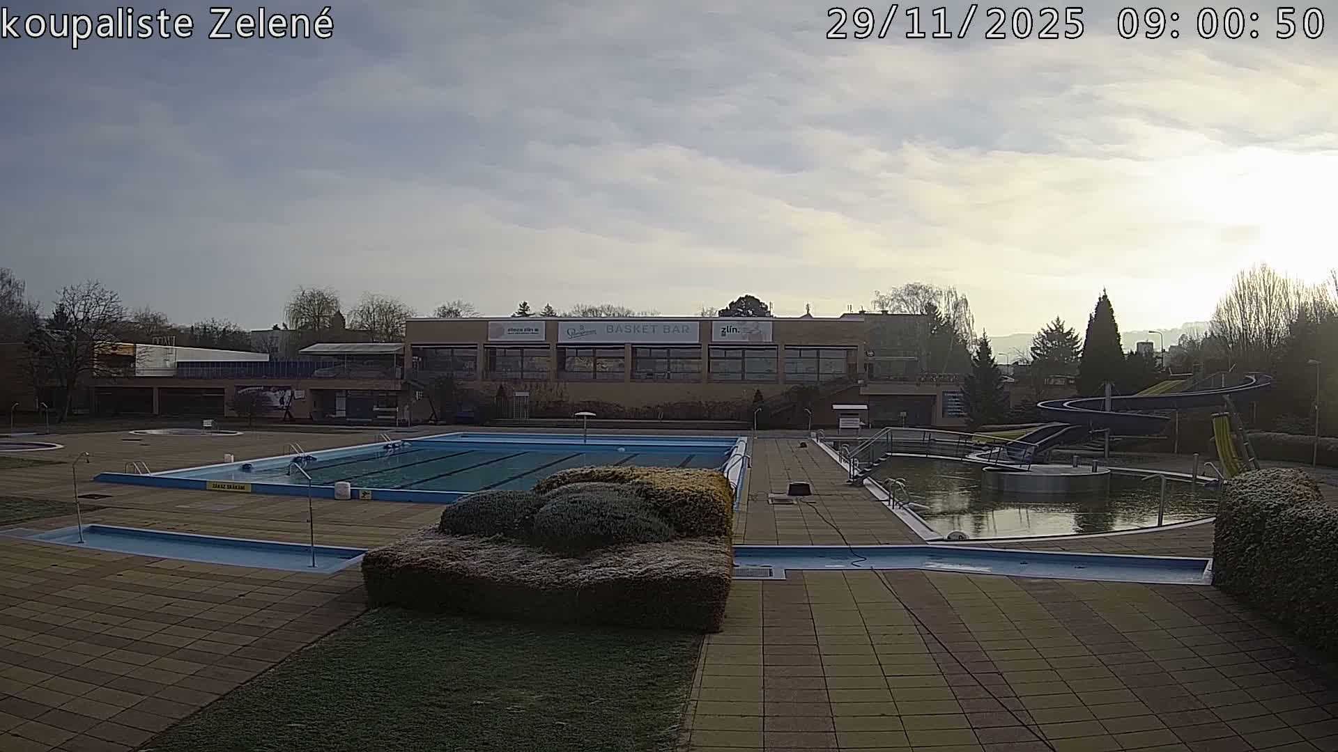 An empty outdoor swimming pool complex, featuring multiple pools and water slides, appears dormant under an overcast sky with patches of frost visible on the surrounding bushes and ground, indicating a chilly morning.