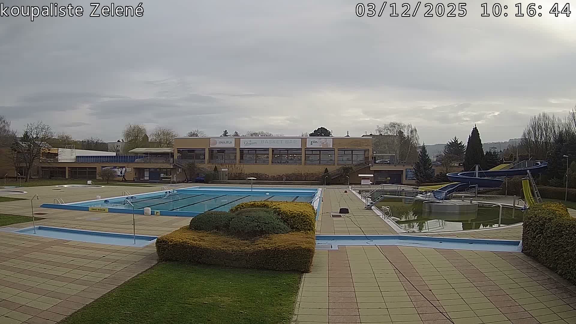 An empty outdoor swimming pool complex, featuring multiple pools (one appearing murky green), water slides, and a facility building, is visible under a uniformly cloudy sky.