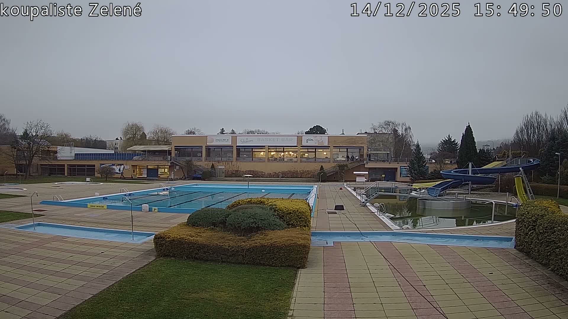 An empty outdoor swimming pool complex, featuring multiple pools (one appearing murky green), water slides, and a facility building, is visible under a uniformly cloudy sky.
