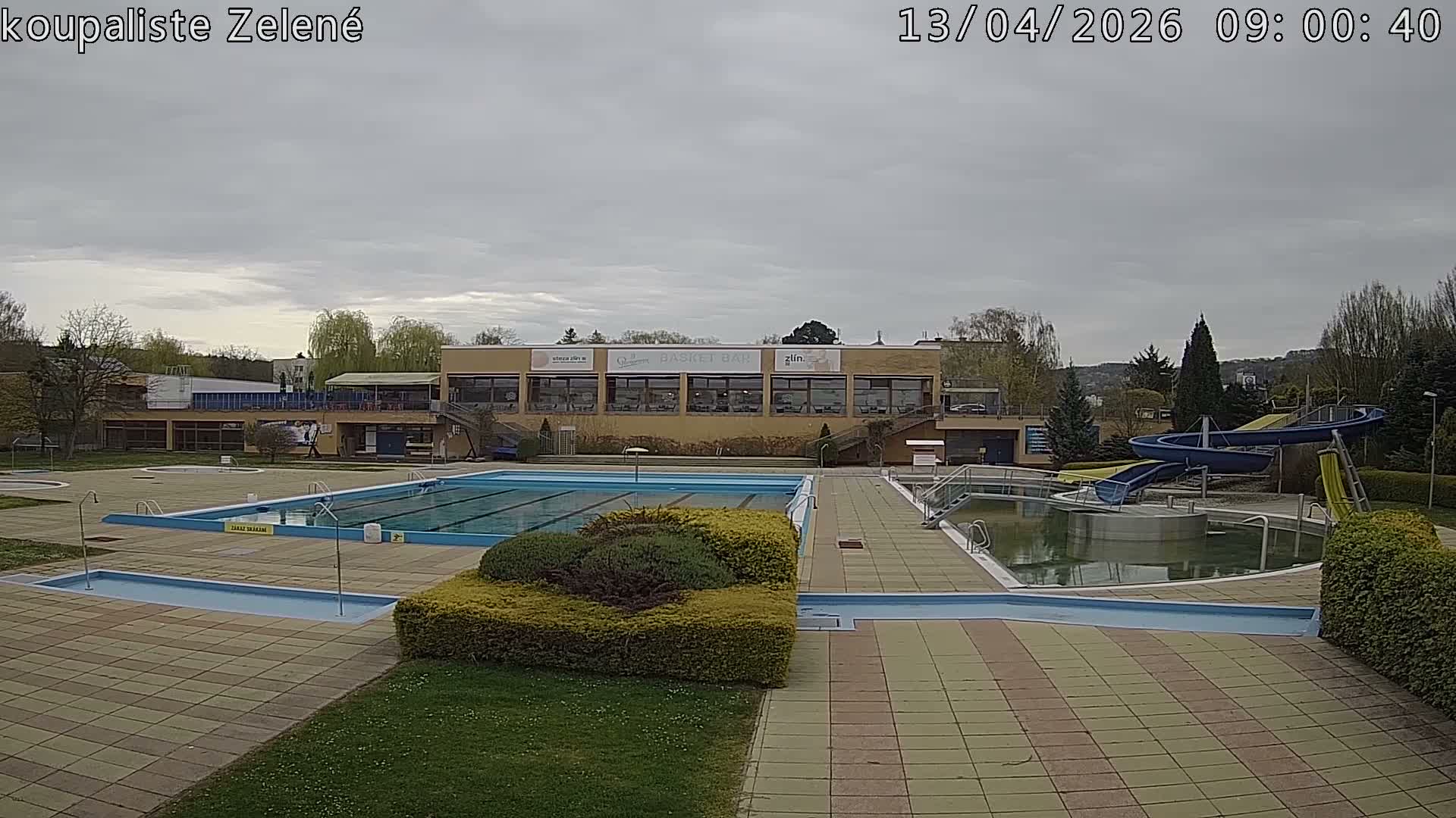 An empty outdoor swimming pool complex, featuring multiple pools (one appearing murky green), water slides, and a facility building, is visible under a uniformly cloudy sky.