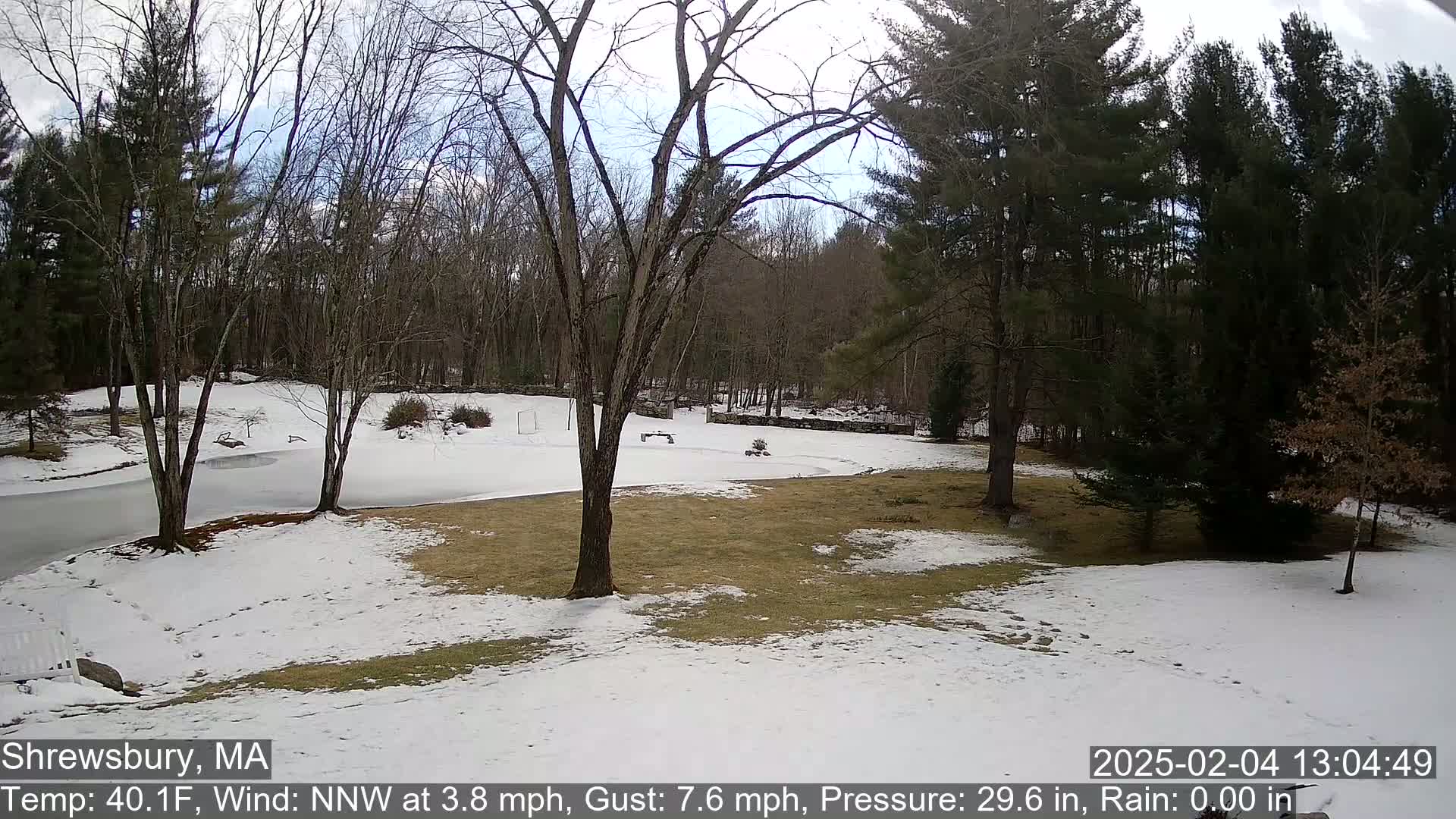 A partially snow-covered yard with bare trees and a partially frozen pond is visible under a partly cloudy sky.