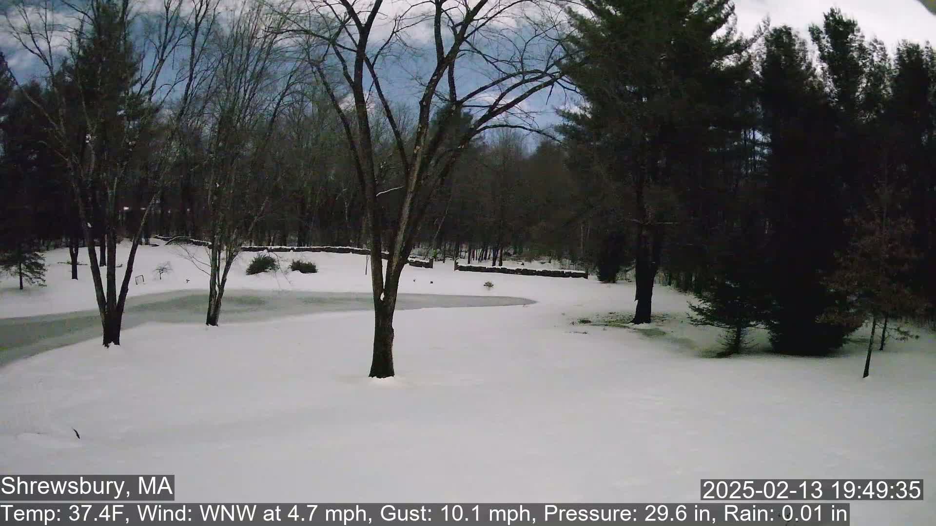 A snow-covered landscape with bare trees and a partially frozen pond under a partly cloudy sky.