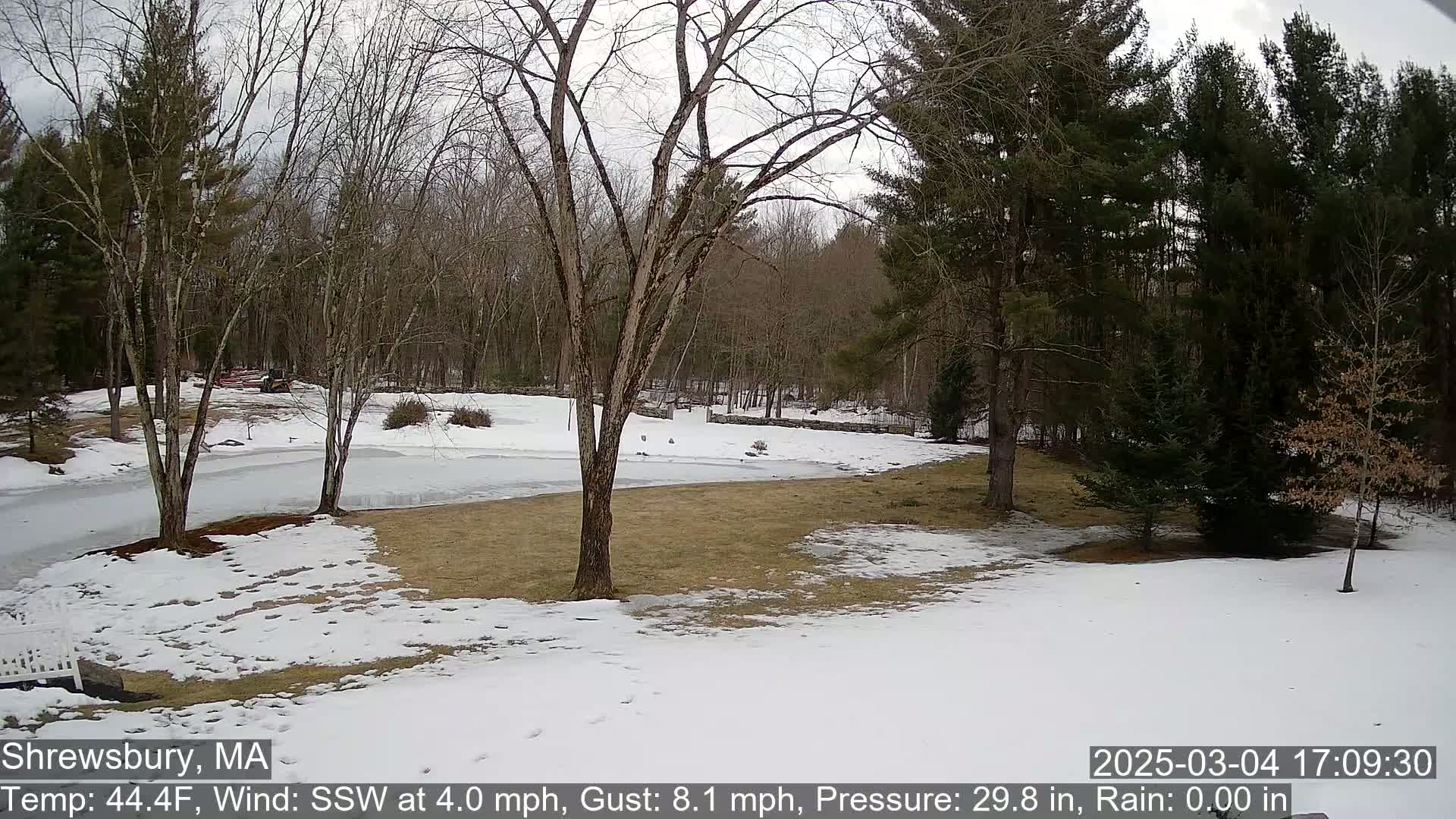 A snow-covered yard with bare trees and a partially frozen body of water in the background under a cloudy sky.