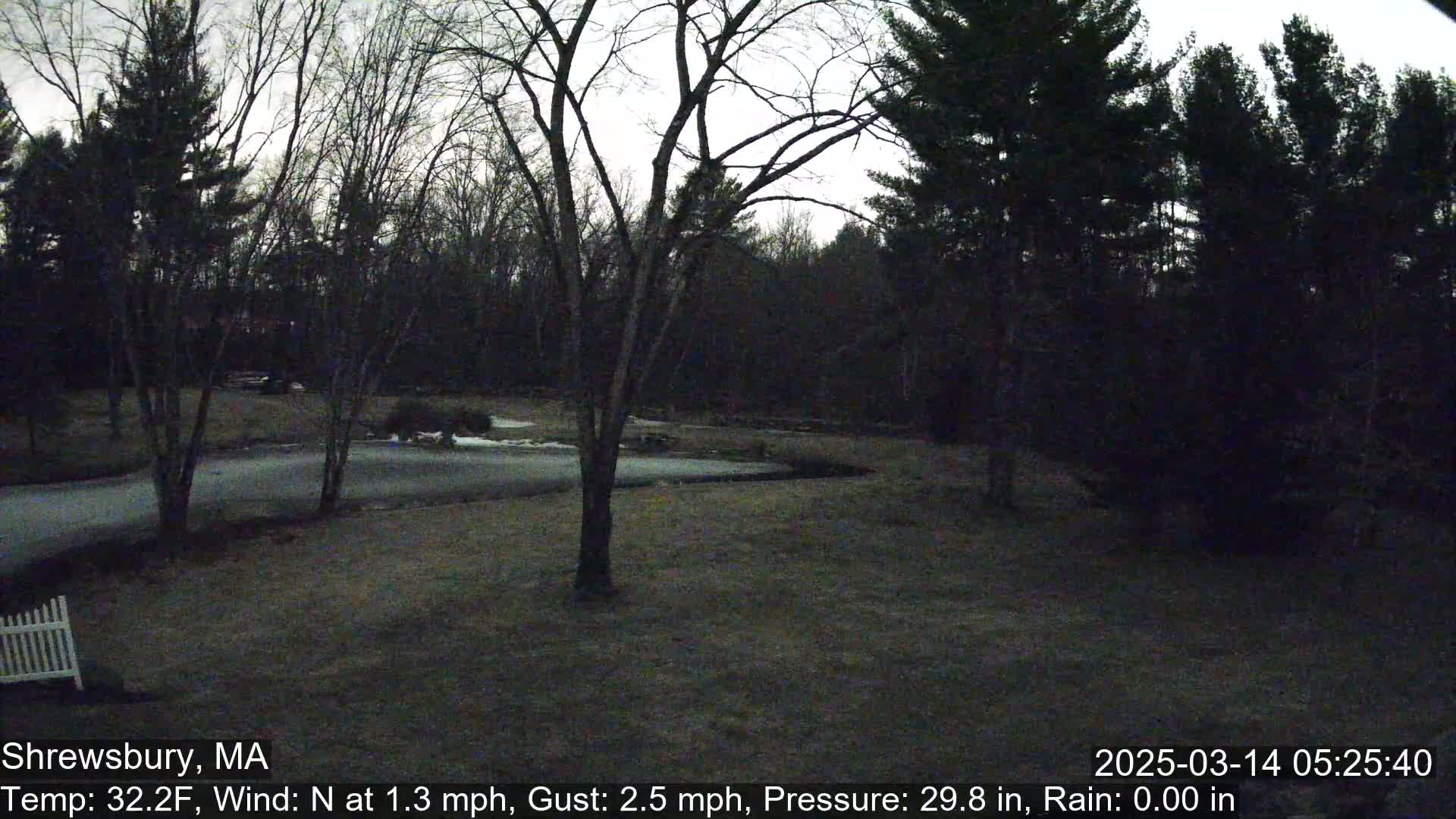 A dimly lit, overcast scene shows bare trees surrounding a grassy yard with a paved driveway, a small body of water partially frozen, and a white picket fence partially visible in the lower left corner.