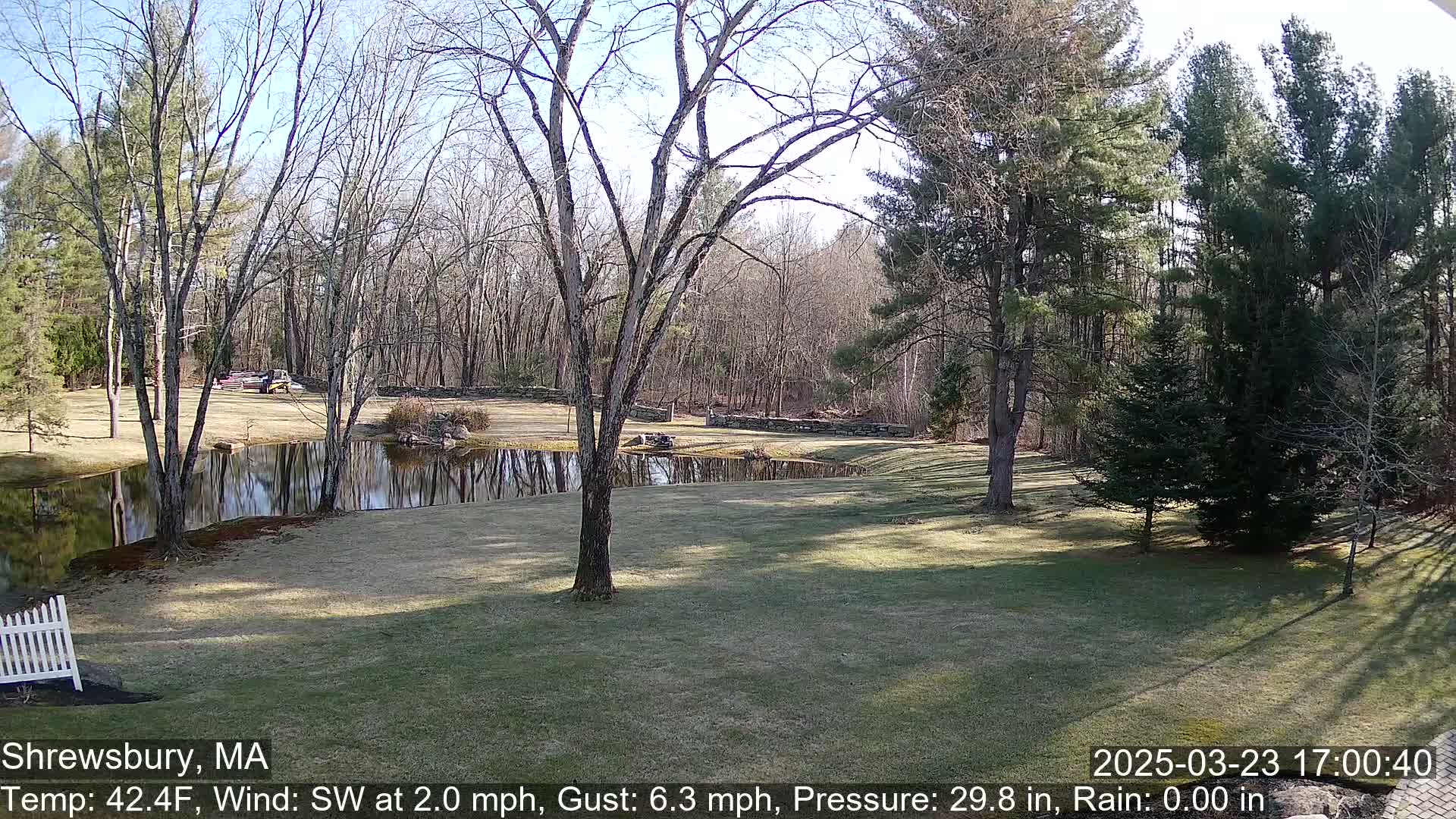 A calm, sunny day reveals a pond reflecting leafless trees and a grassy lawn bordered by a stone wall and evergreen trees, with a white picket fence partially visible in the bottom left corner.