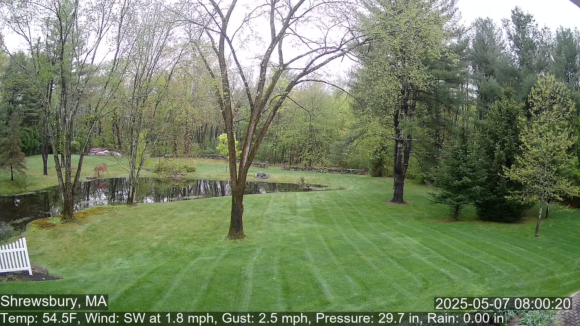 A lush green lawn stretches towards a pond bordered by trees and stone walls under an overcast sky.