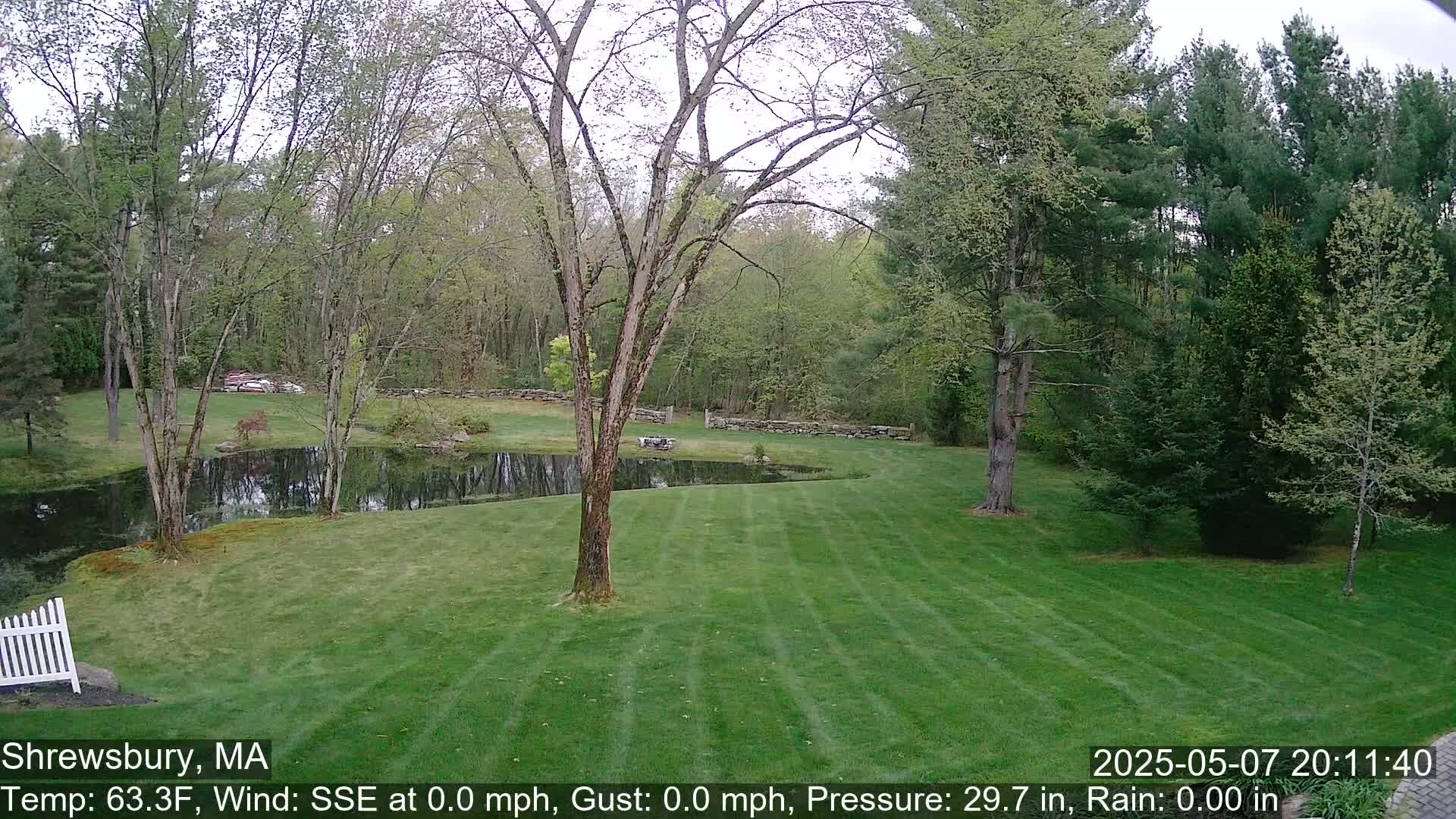 A neatly mown lawn stretches from a white picket fence toward a calm pond nestled among leafy green trees under a partly cloudy sky.