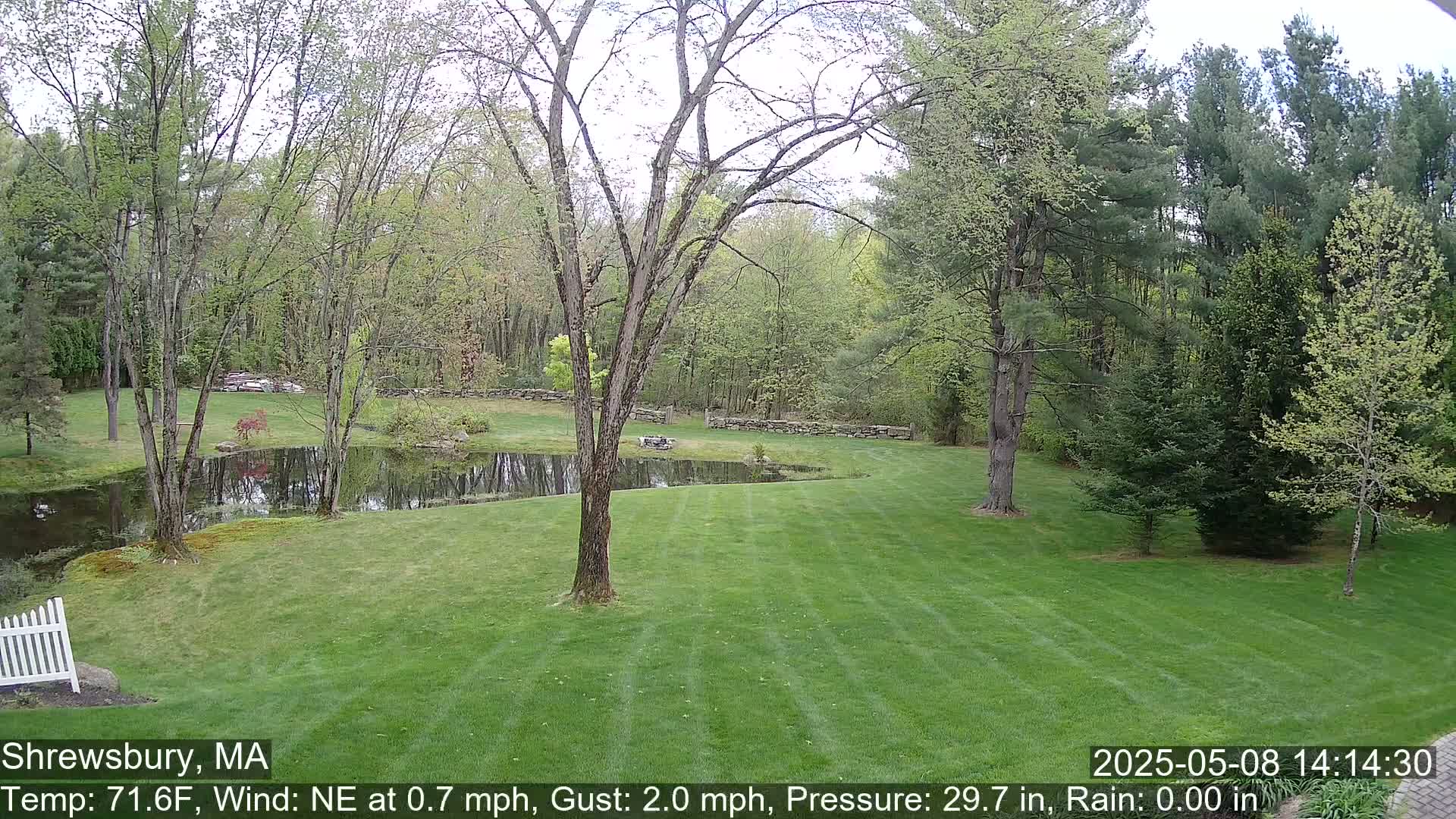 A large, manicured lawn stretches toward a pond and stone wall, surrounded by deciduous trees under a partly cloudy sky.