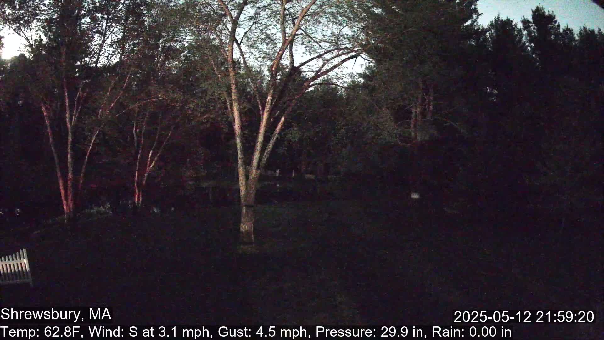A dark, nighttime scene shows a yard with trees and a portion of a white fence, under a clear sky with a light south wind.