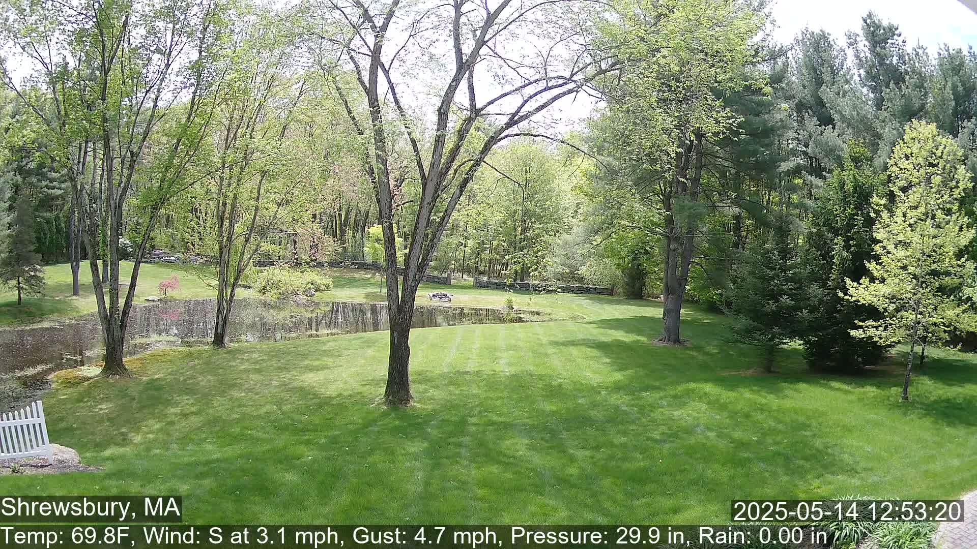 A lush green lawn stretches out before a pond, surrounded by deciduous trees under a partly sunny sky.
