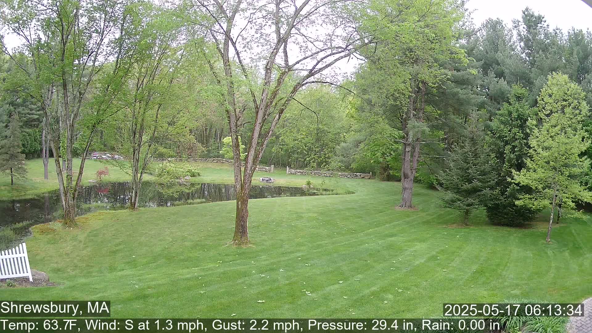 A green lawn slopes down to a small pond surrounded by trees and a stone wall, under a mostly cloudy sky.
