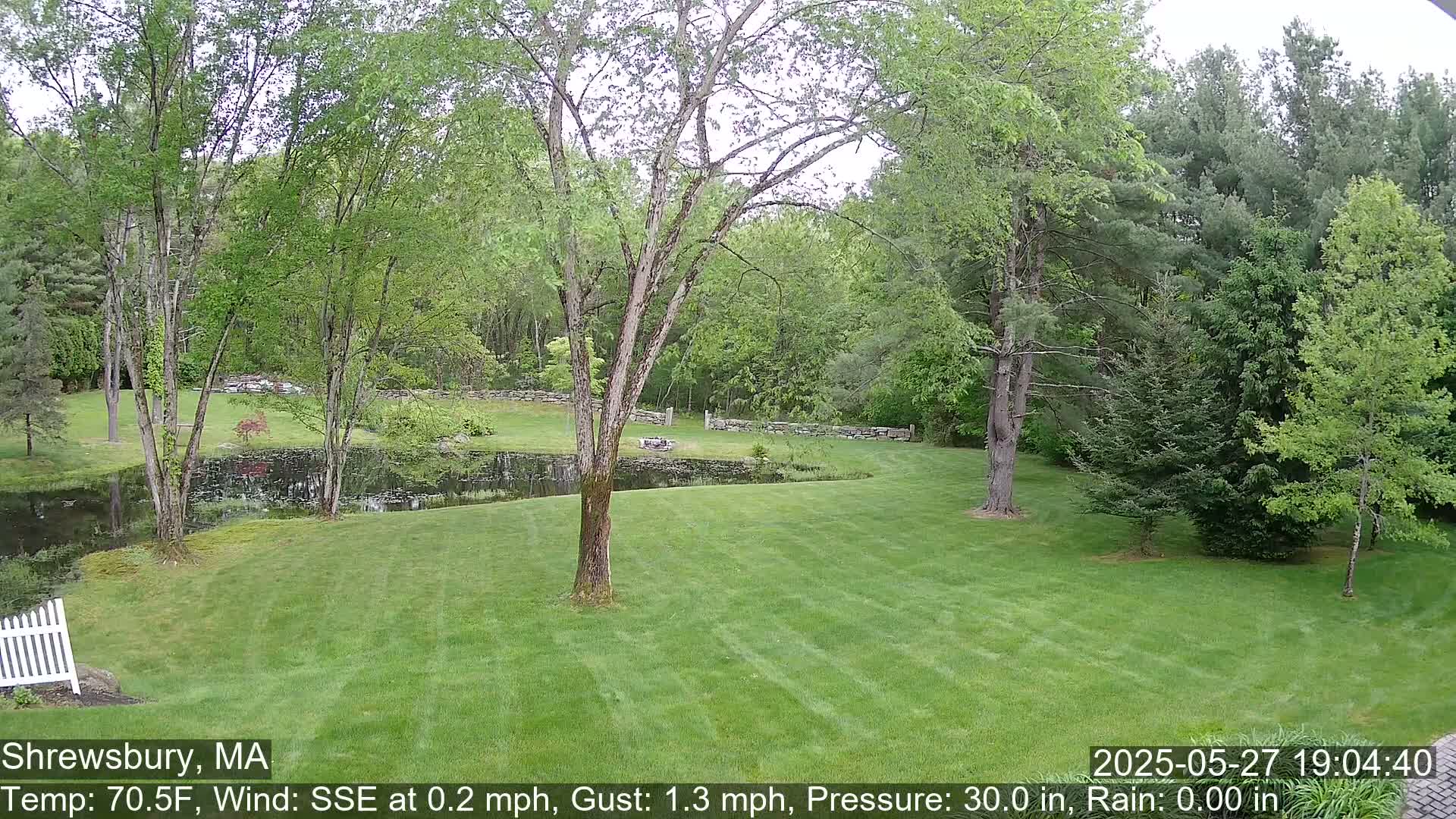A green lawn slopes down to a pond surrounded by trees and stone walls under a cloudy sky.