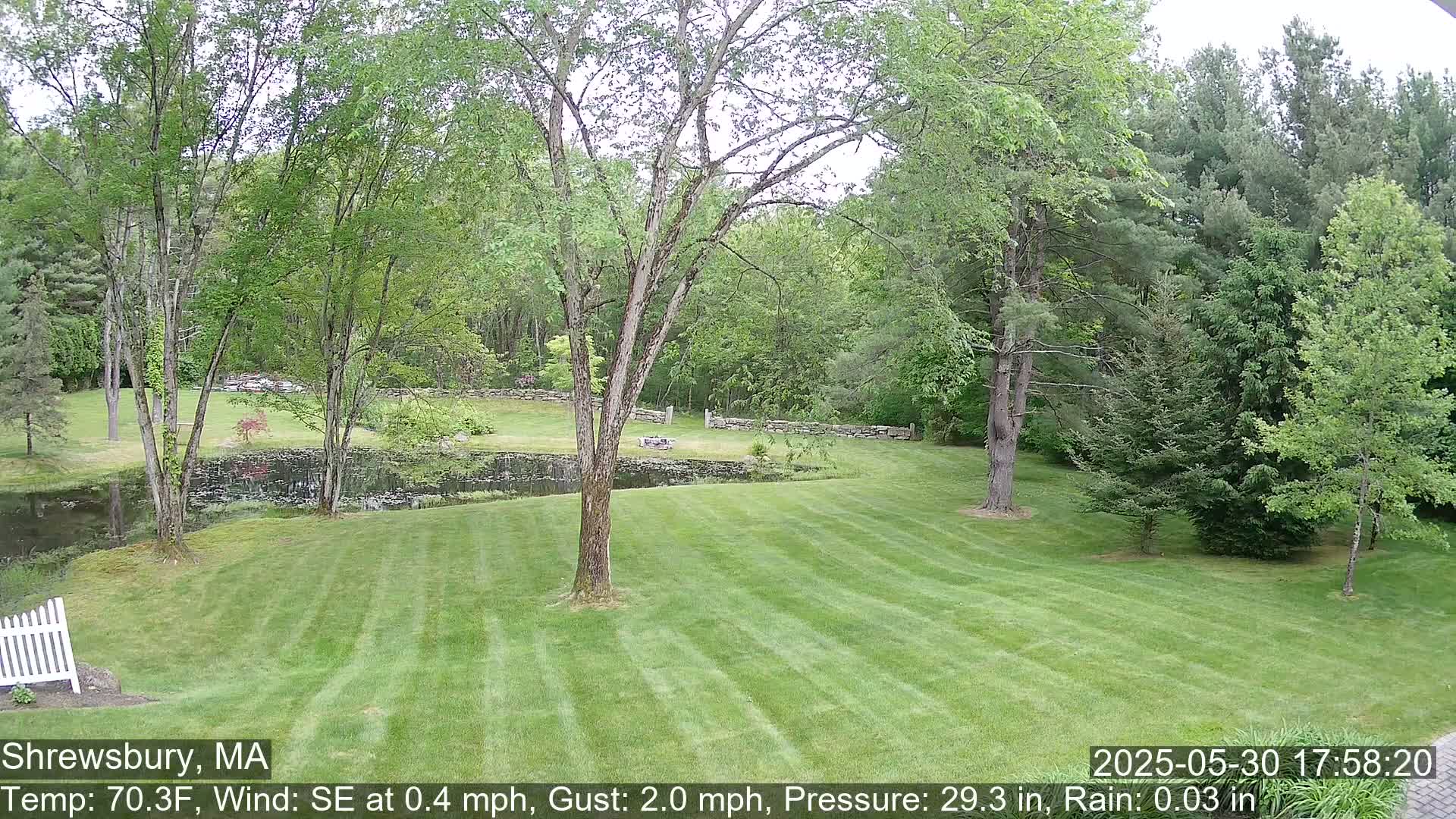 A neatly mown lawn slopes gently down to a small pond, surrounded by lush green trees and stone walls under a lightly rainy sky.