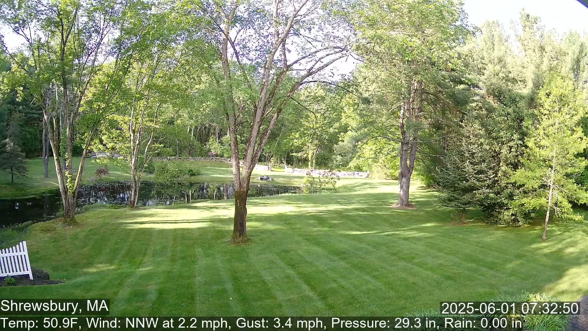 A neatly mown lawn slopes gently down toward a pond nestled amongst leafy green trees on a sunny day with a light north-northwest wind.