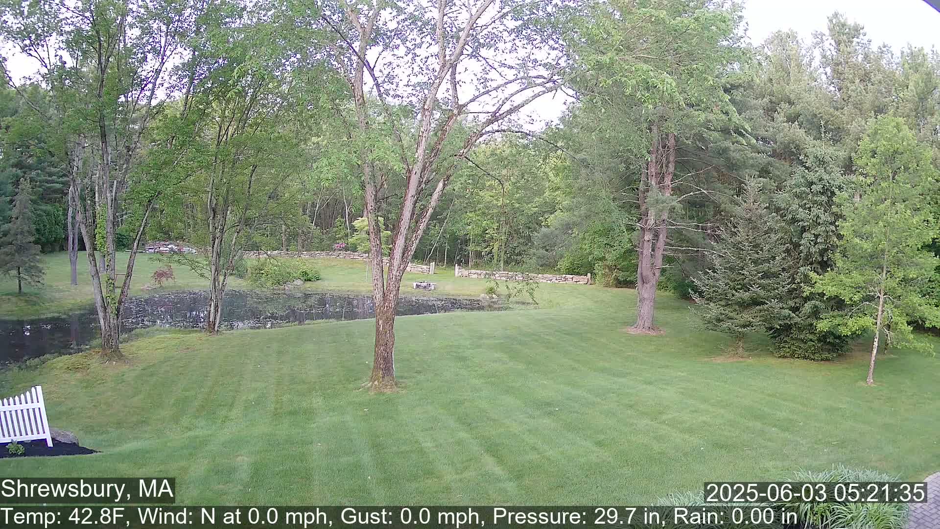 A grassy lawn extends to a calm pond bordered by a stone wall and lush green trees under a cloudy sky.