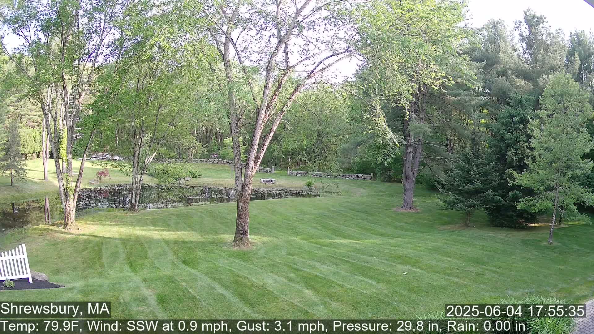 A neatly mown lawn stretches towards a pond bordered by stone walls and lush green trees under a partly sunny sky.