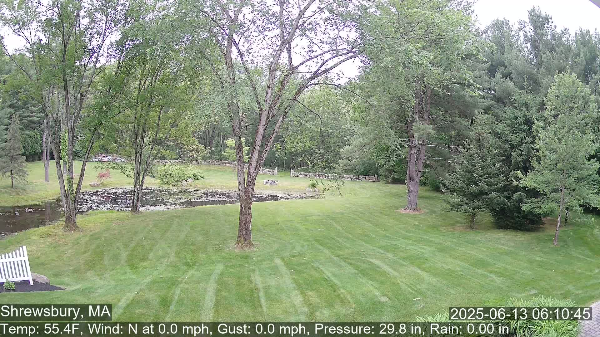 A green lawn stretches toward a pond and stone wall, surrounded by lush green trees under an overcast sky.