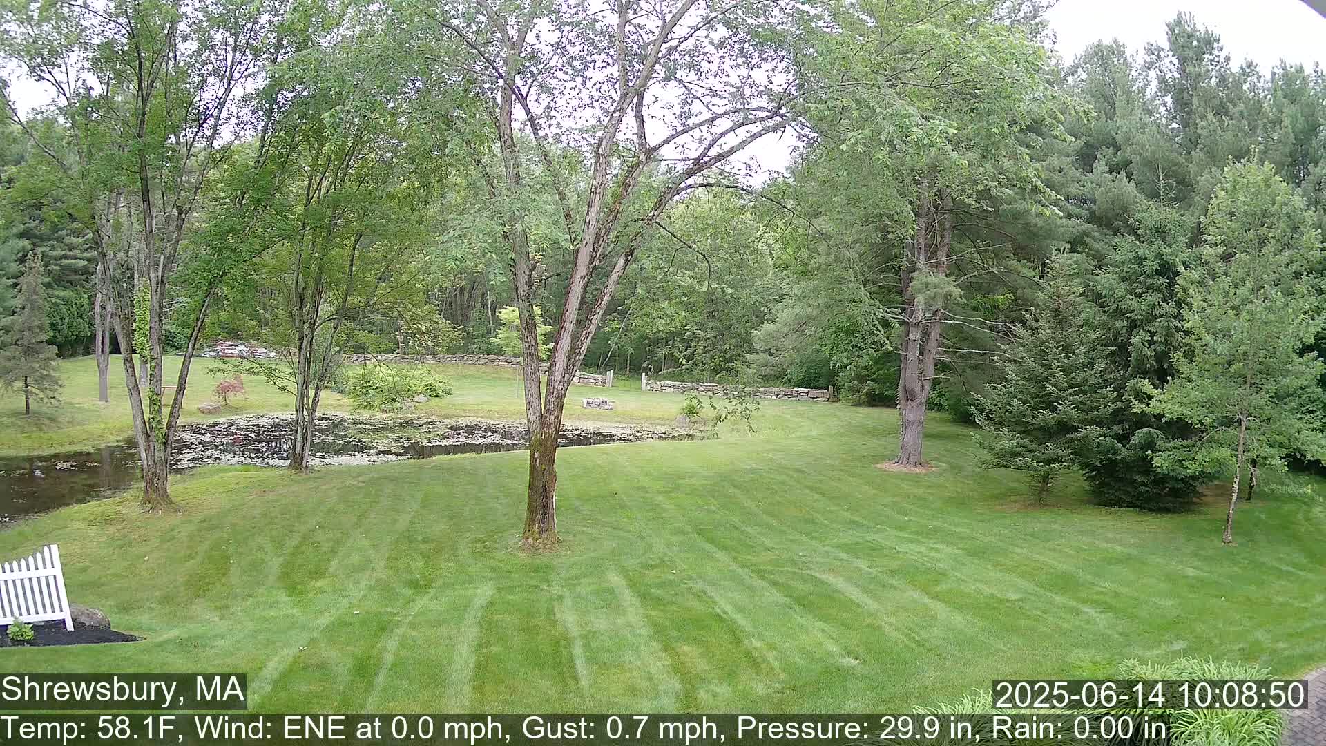 A verdant lawn stretches toward a pond and stone wall, bordered by lush green trees under a partly cloudy sky.
