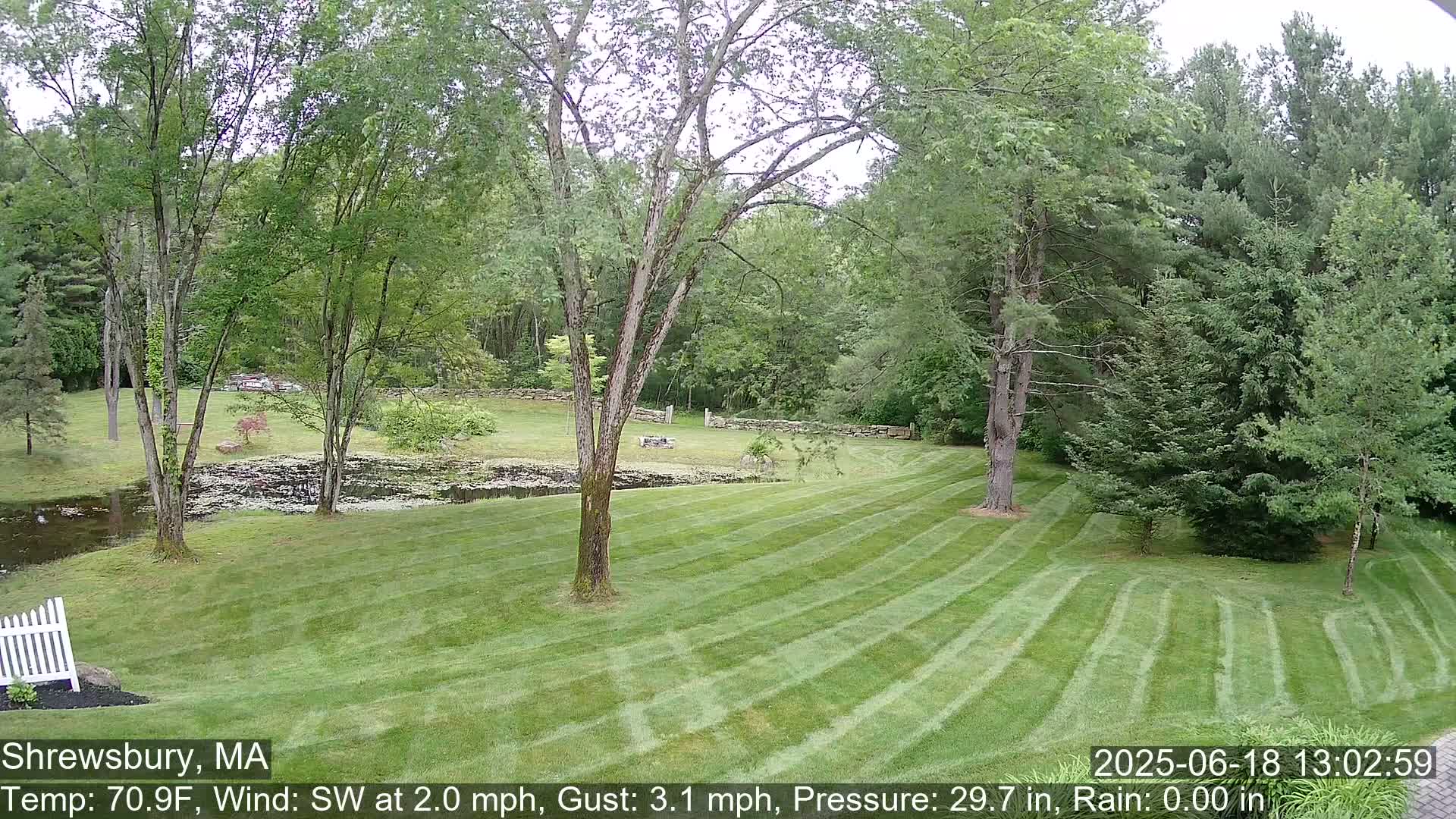 A freshly mown lawn slopes down to a small pond, surrounded by lush green trees under a partly sunny sky.