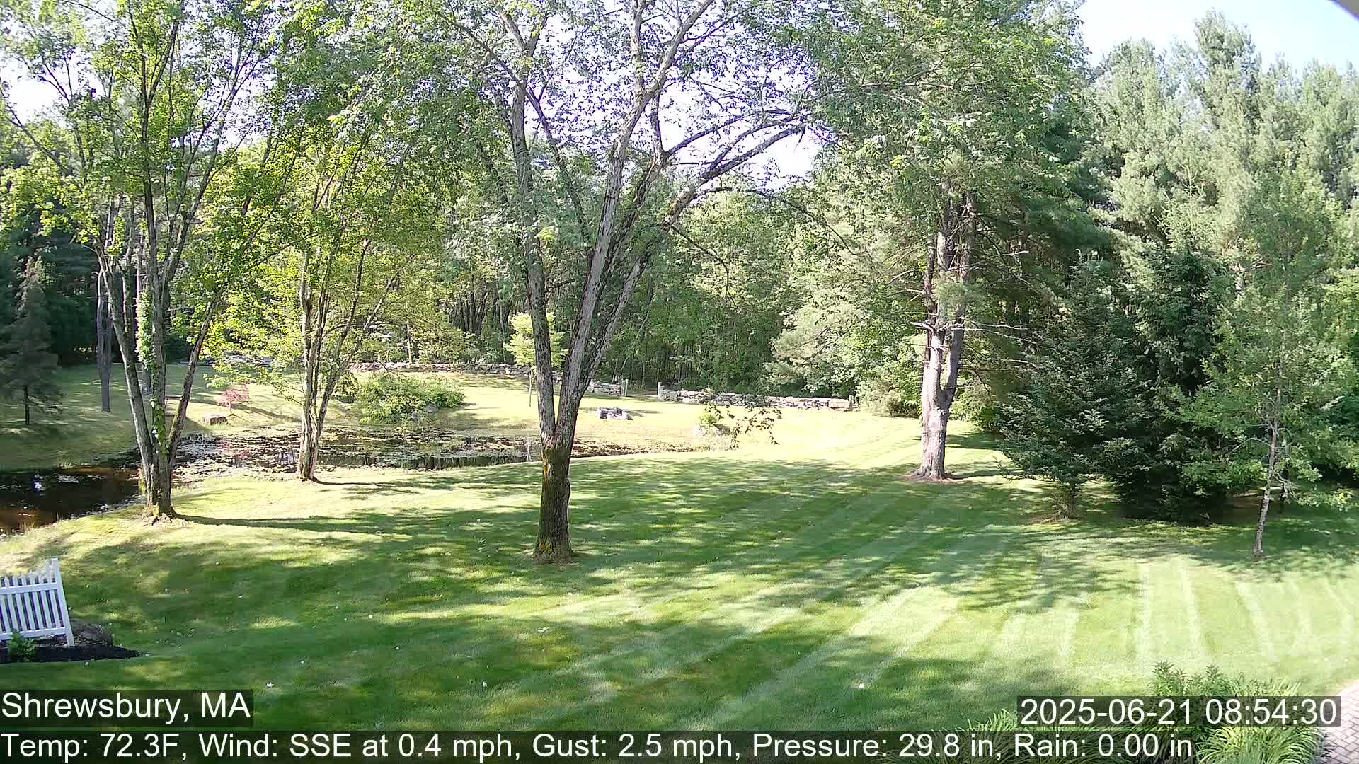 A sunny day reveals a neatly mown lawn bordered by trees and a small pond, beyond a stone wall and more trees.